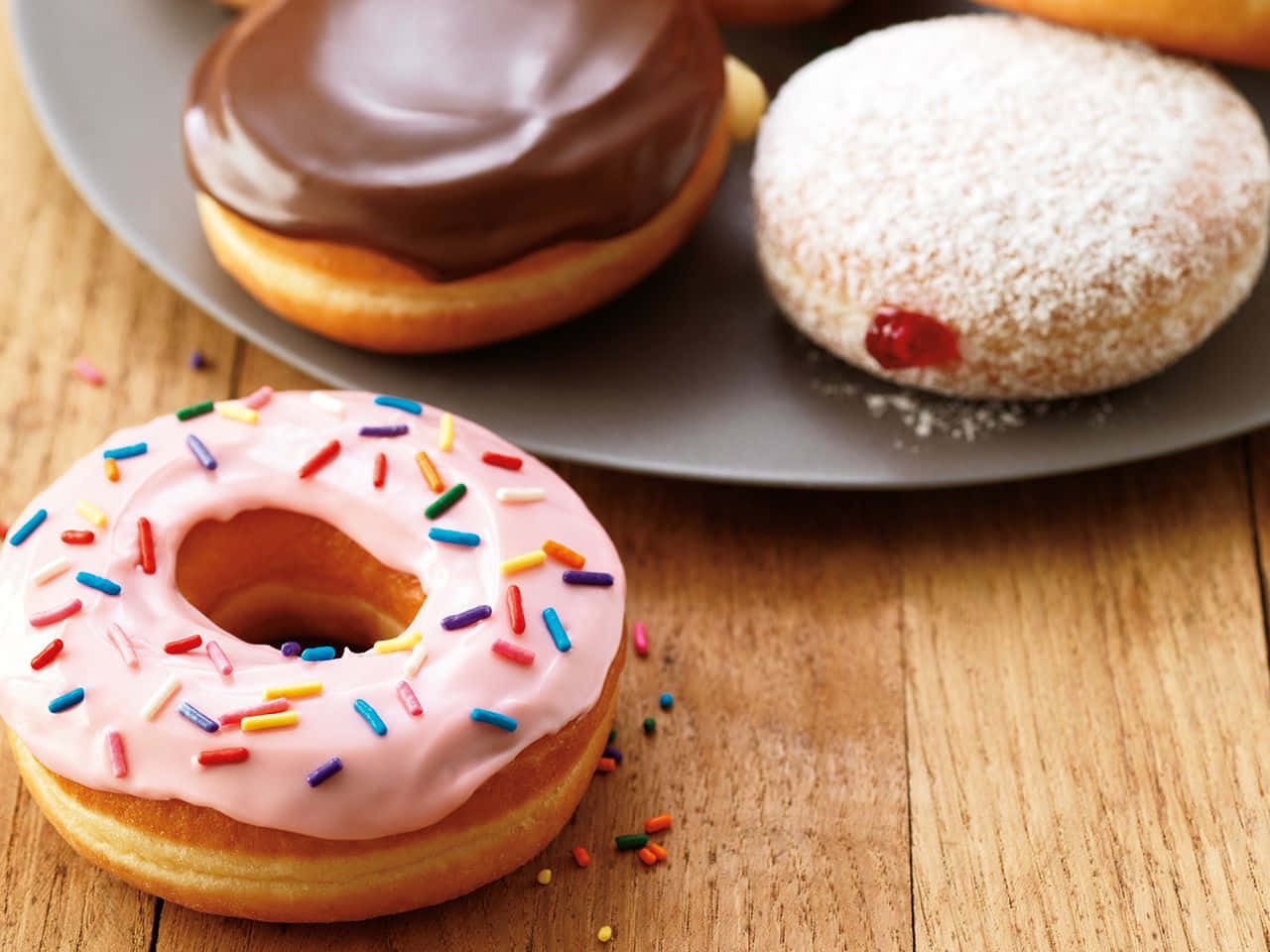 Dunkin Donuts 桌布 壁紙 — 三個美味的甜甜圈，上面有粉紅色糖粒、巧克力糖霜和糖粉，誘人地放在木桌上。