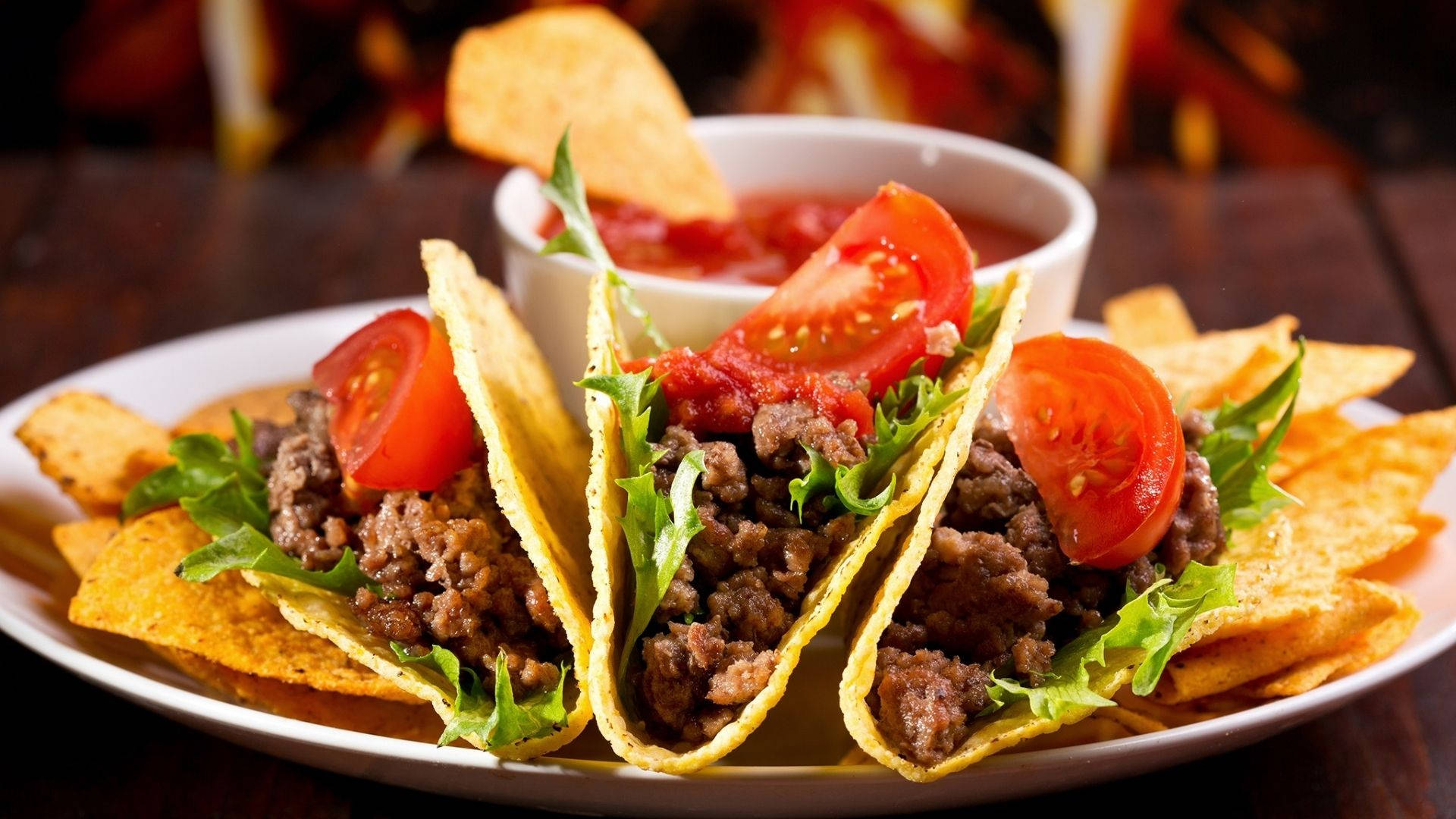 Taco 桌布 壁紙 — 三個脆皮墨西哥捲餅，內餡為調味牛絞肉、新鮮生菜和番茄片，放在一個被玉米片和一碗莎莎醬環繞的盤子上。