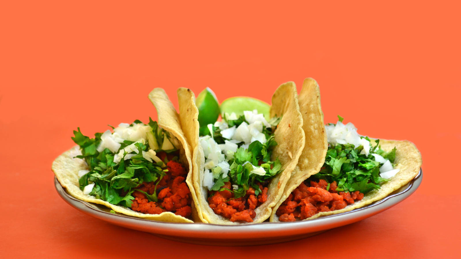 Tacos 桌布 壁紙 — 三個脆皮墨西哥捲餅，餡料有紅肉、香菜和洋蔥，放在盤子上，背景是鮮豔的橙色。