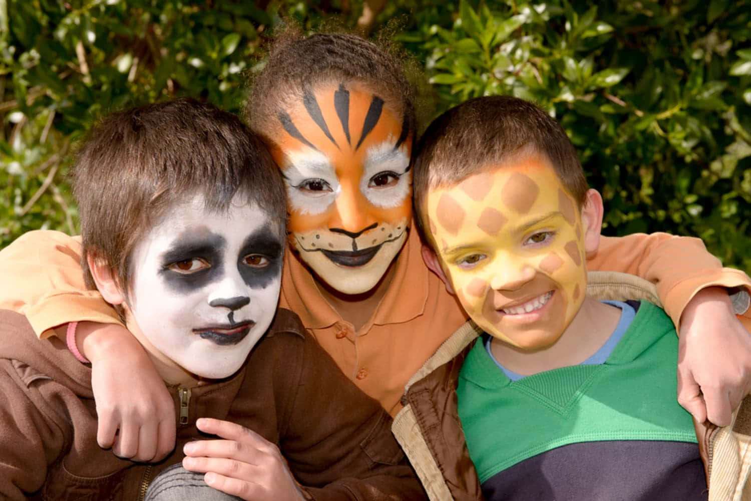 Halloween Face Paint 桌布 壁紙 — 三個臉上畫著動物圖案的孩子在綠色樹葉背景下開心地擁抱。