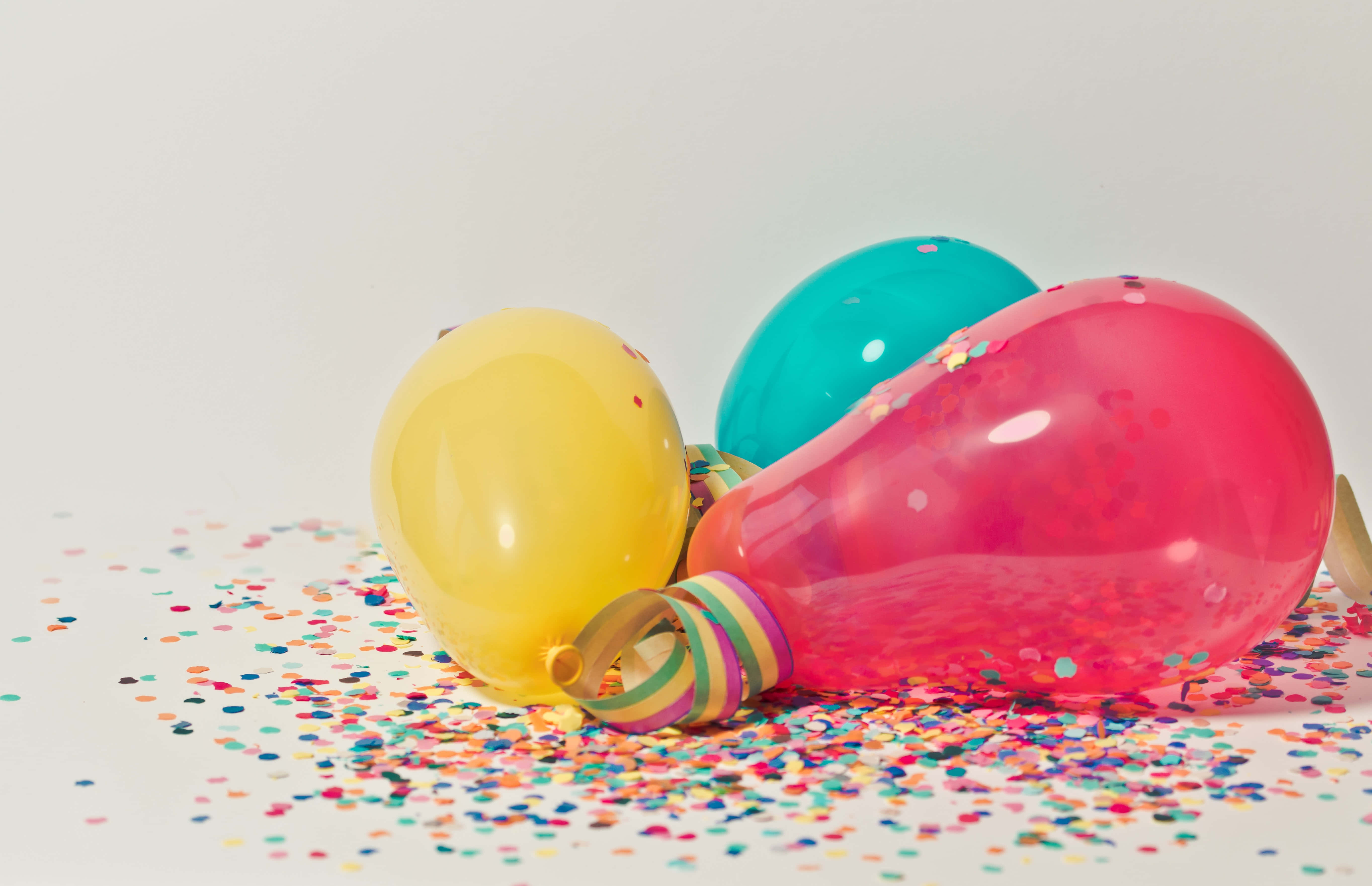 Balloon 桌布 壁紙 — 三個色彩繽紛、裝滿五彩紙屑的氣球放在白色表面上，周圍散落著彩虹亮片。