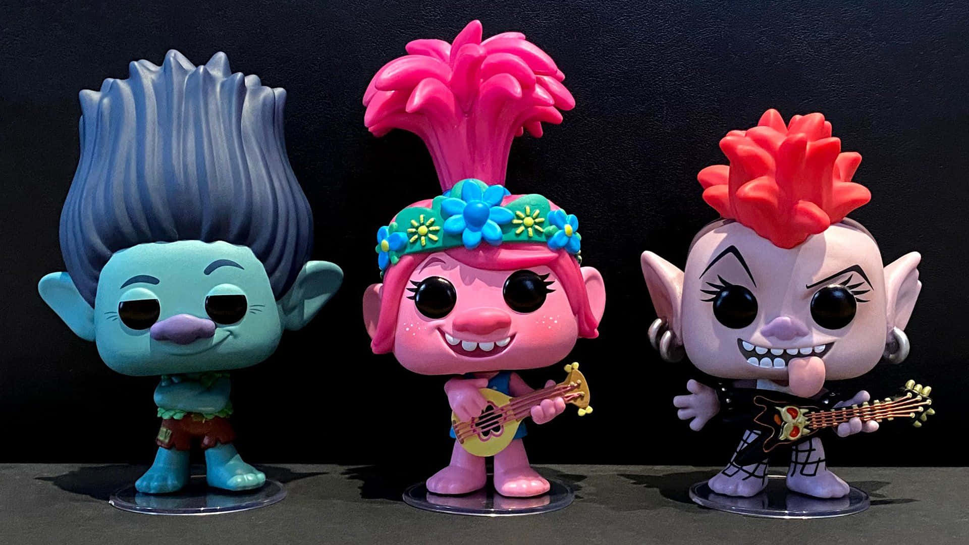 魔髮精靈 桌布 壁紙 — 三個色彩繽紛的 Trolls 角色 Funko Pop! 人偶在深色背景前拿著吉他擺姿勢。