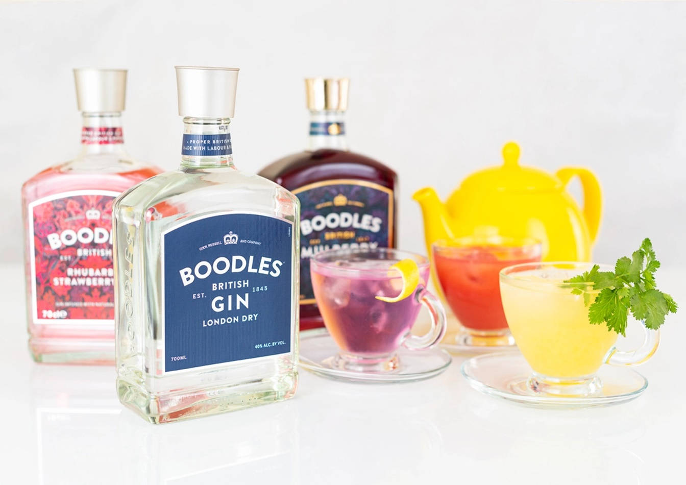 cocktail 桌布 壁紙 — 三個色彩繽紛的Boodles琴酒瓶和充滿活力的雞尾酒，置於明亮的白色反光表面上。