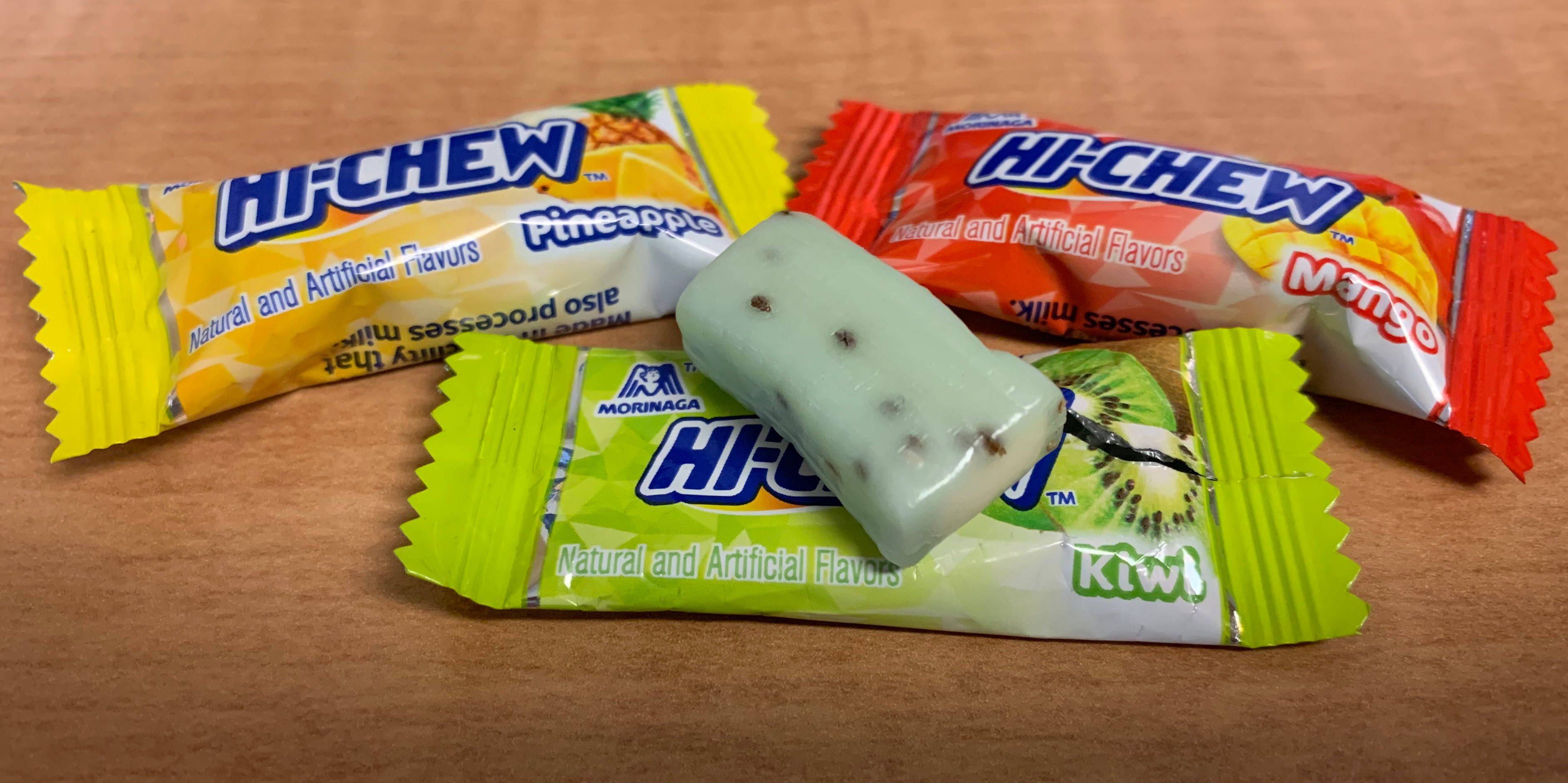 chewy 桌布 壁紙 — 三個色彩繽紛的Hi-Chew糖果包裝紙和一個已拆開包裝的奇異果味軟糖放在木質表面上。