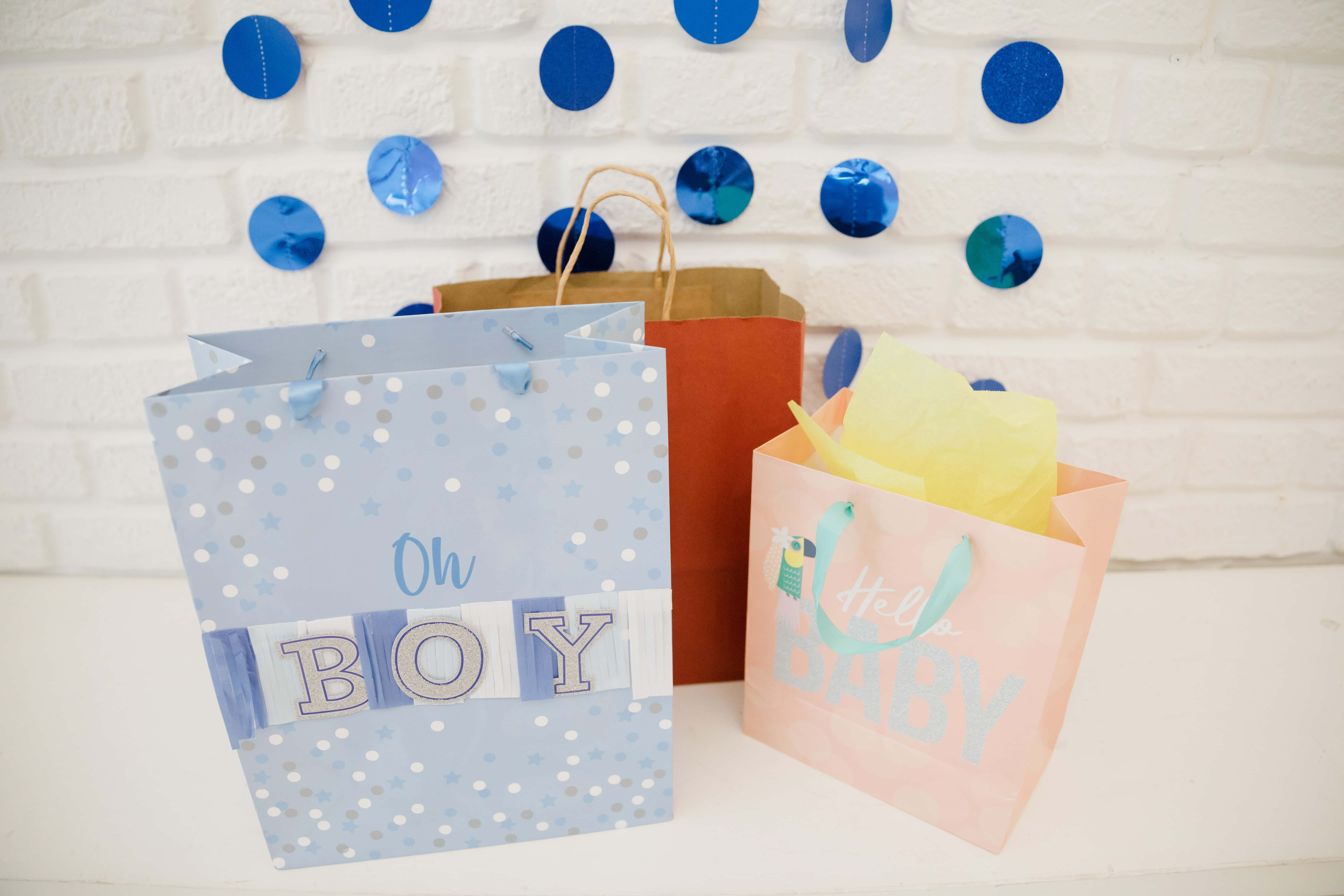 Party 桌布 壁紙 — 三個色彩繽紛的嬰兒派對禮物袋，上面有「Oh BOY」和「Hello BABY」字樣，放在一面飾有藍色圓形裝飾的白色磚牆前。