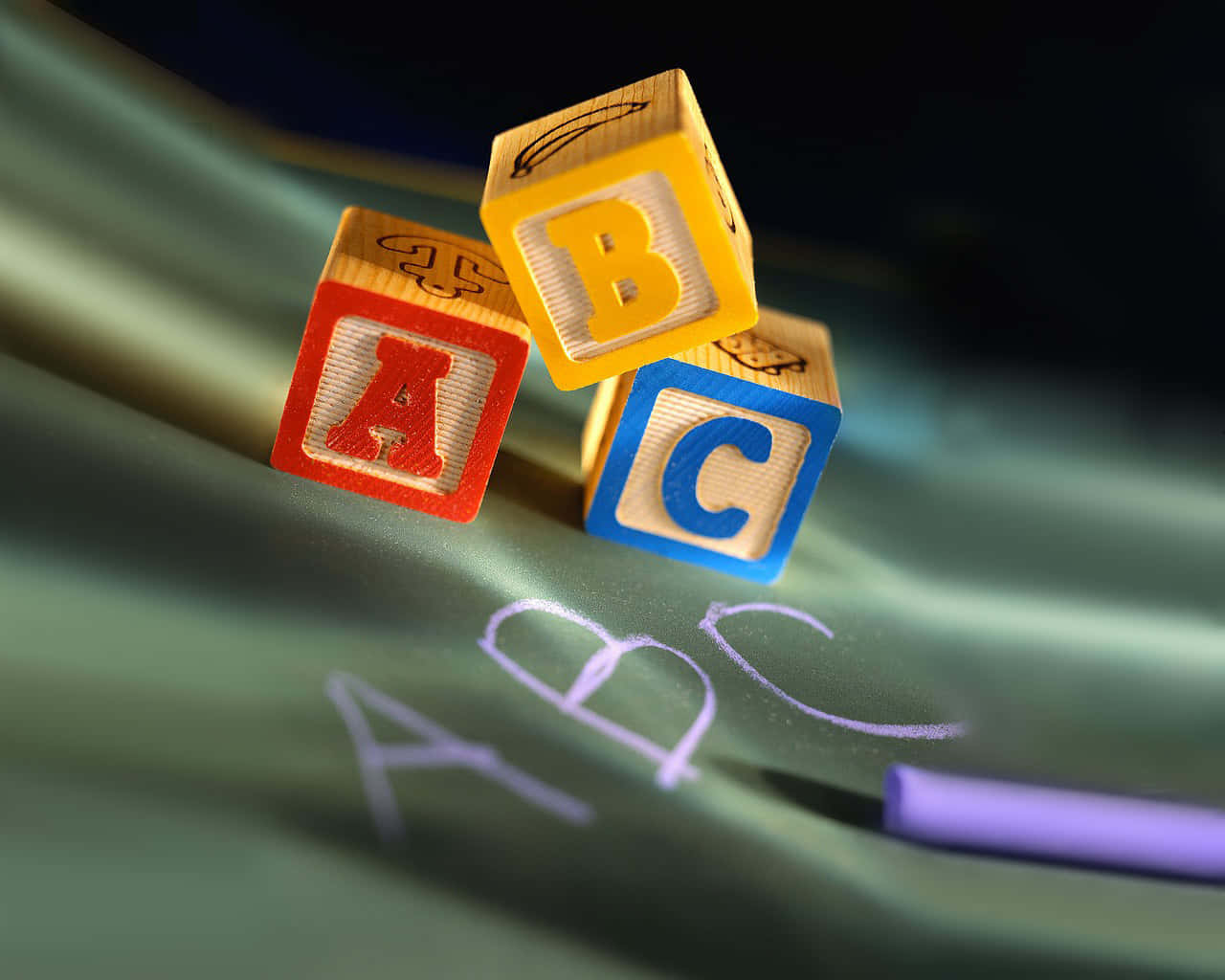 abc 桌布 壁紙 — 三個色彩繽紛的木製字母積木拼出ABC，放在綠色布料上，布料上有粉筆畫出的字母，附近還有一支紫色粉筆。