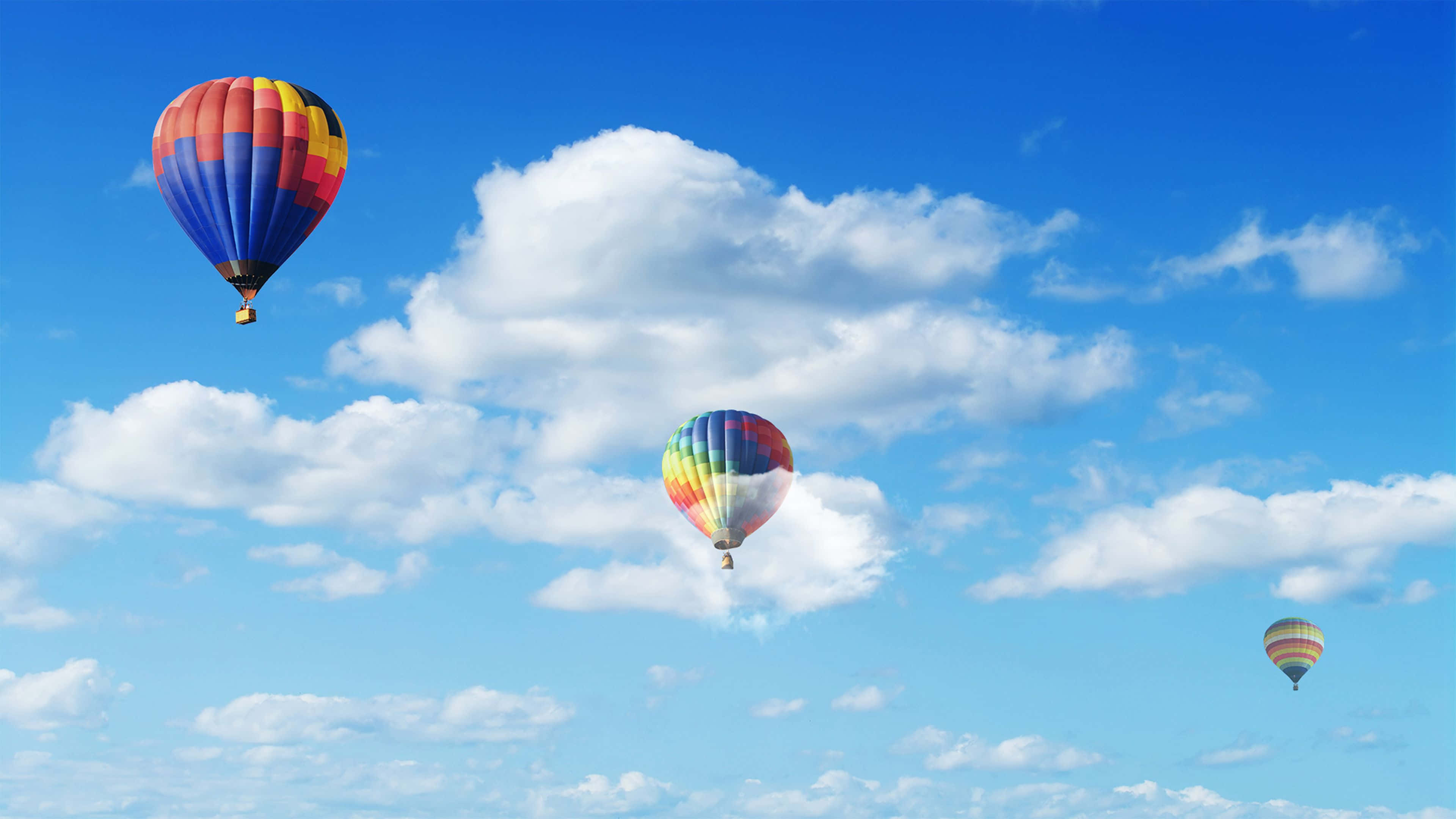 Hot Air Balloon 桌布 壁紙 — 三個色彩繽紛的熱氣球在點綴著蓬鬆白雲的明亮藍天中優雅飄浮。