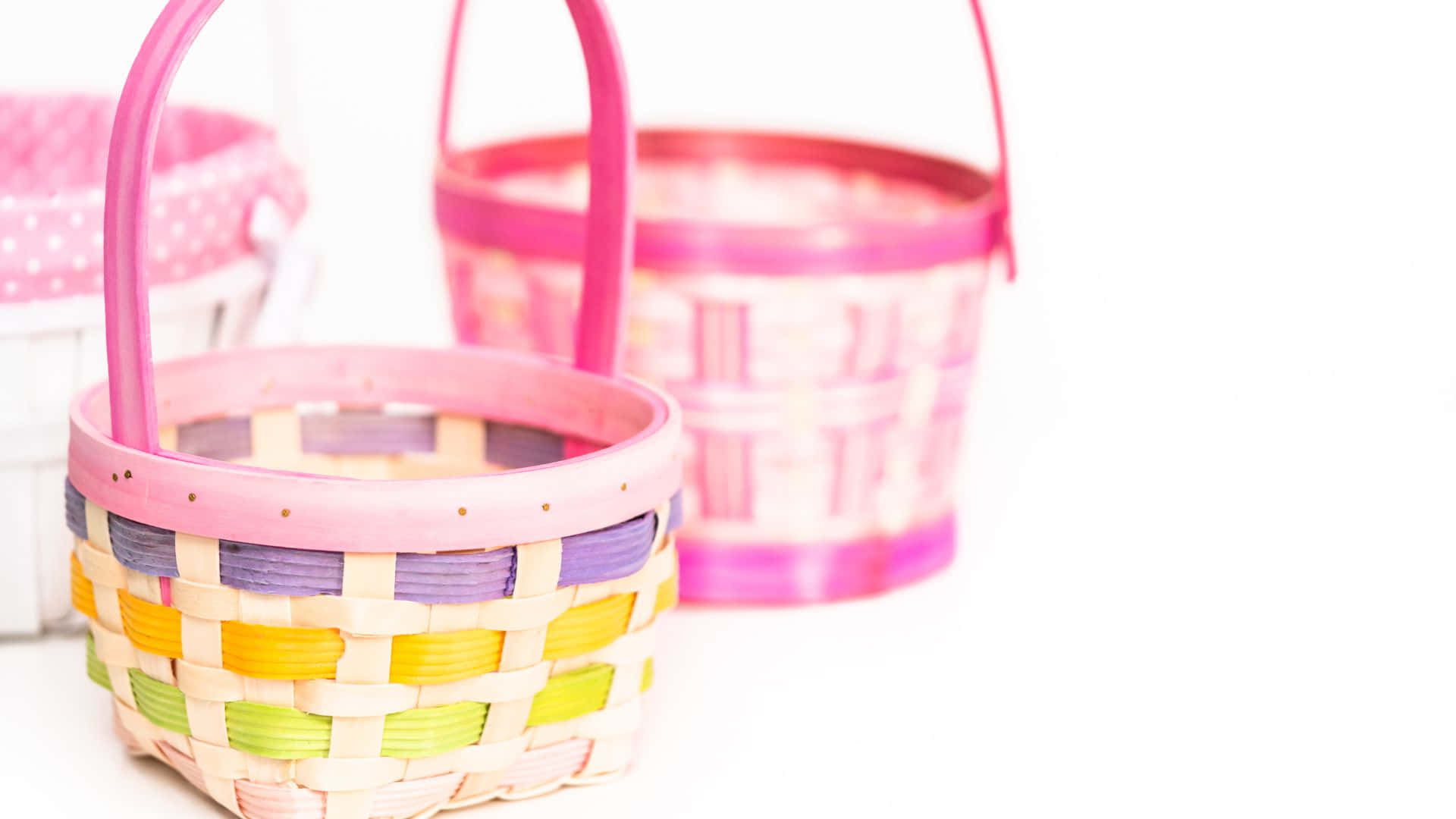 Easter Basket 桌布 壁紙 — 三個色彩繽紛的編織籃子，帶有粉紅色手柄，以淺景深排列在明亮的白色背景上。