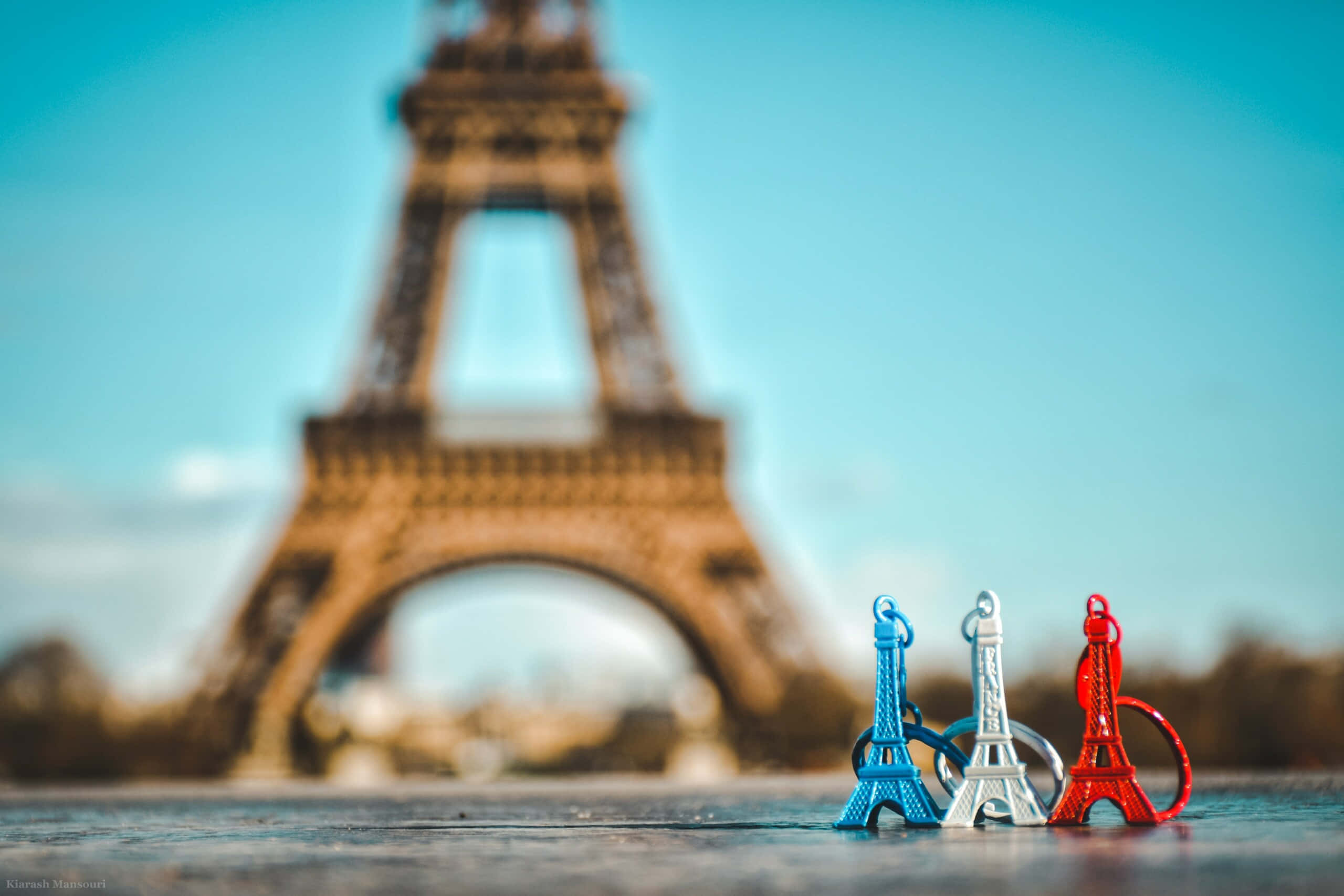 french 桌布 壁紙 — 三個色彩繽紛的艾菲爾鐵塔鑰匙圈在行人路上清晰對焦，真正的艾菲爾鐵塔在背後模糊，襯著清澈的藍天。