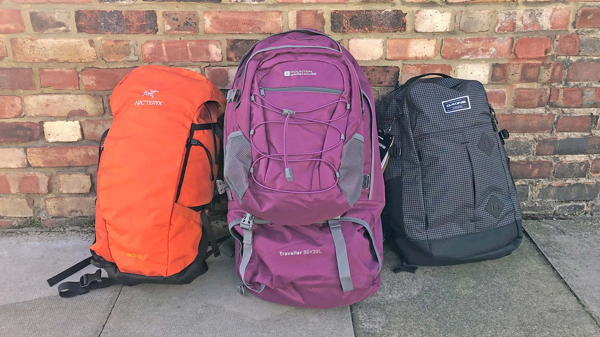 Purple Travel Bag 桌布 壁紙 — 三個色彩繽紛的遠足背包（橙色、紫色和黑色）靠在混凝土人行道上的風化磚牆上。