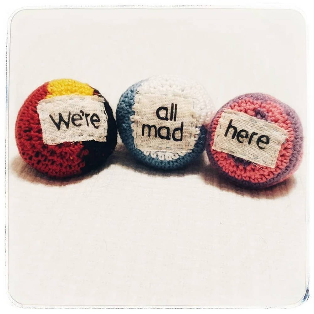 藝術 桌布 壁紙 — 三個色彩繽紛的鉤針編織球，上面有寫著「We're all mad here」的布料標籤，排成一排放在平坦的表面上。