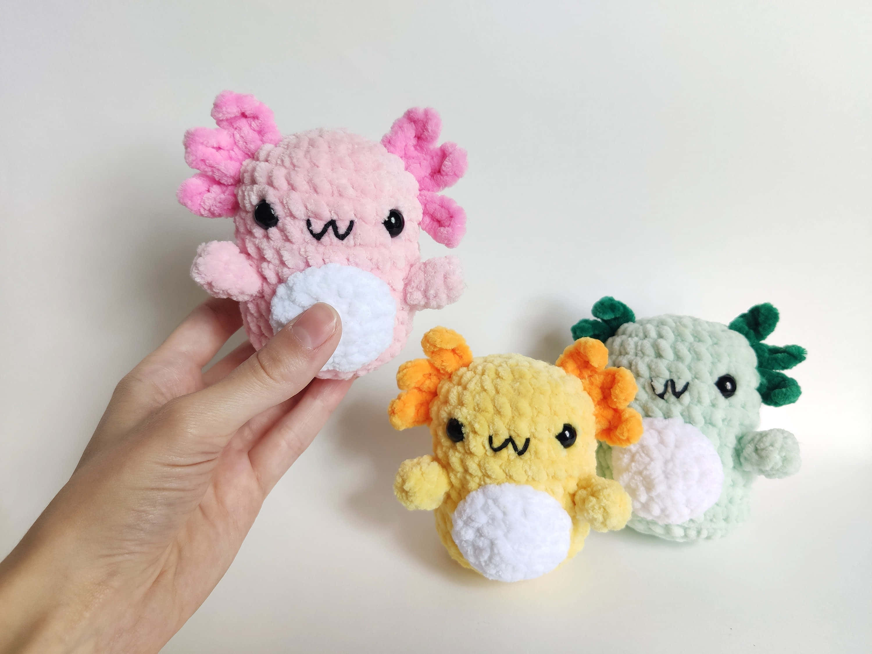 Crochet 桌布 壁紙 — 三個色彩繽紛的鉤針編織蠑螈毛絨玩具，面帶微笑，在純白色背景前展示。