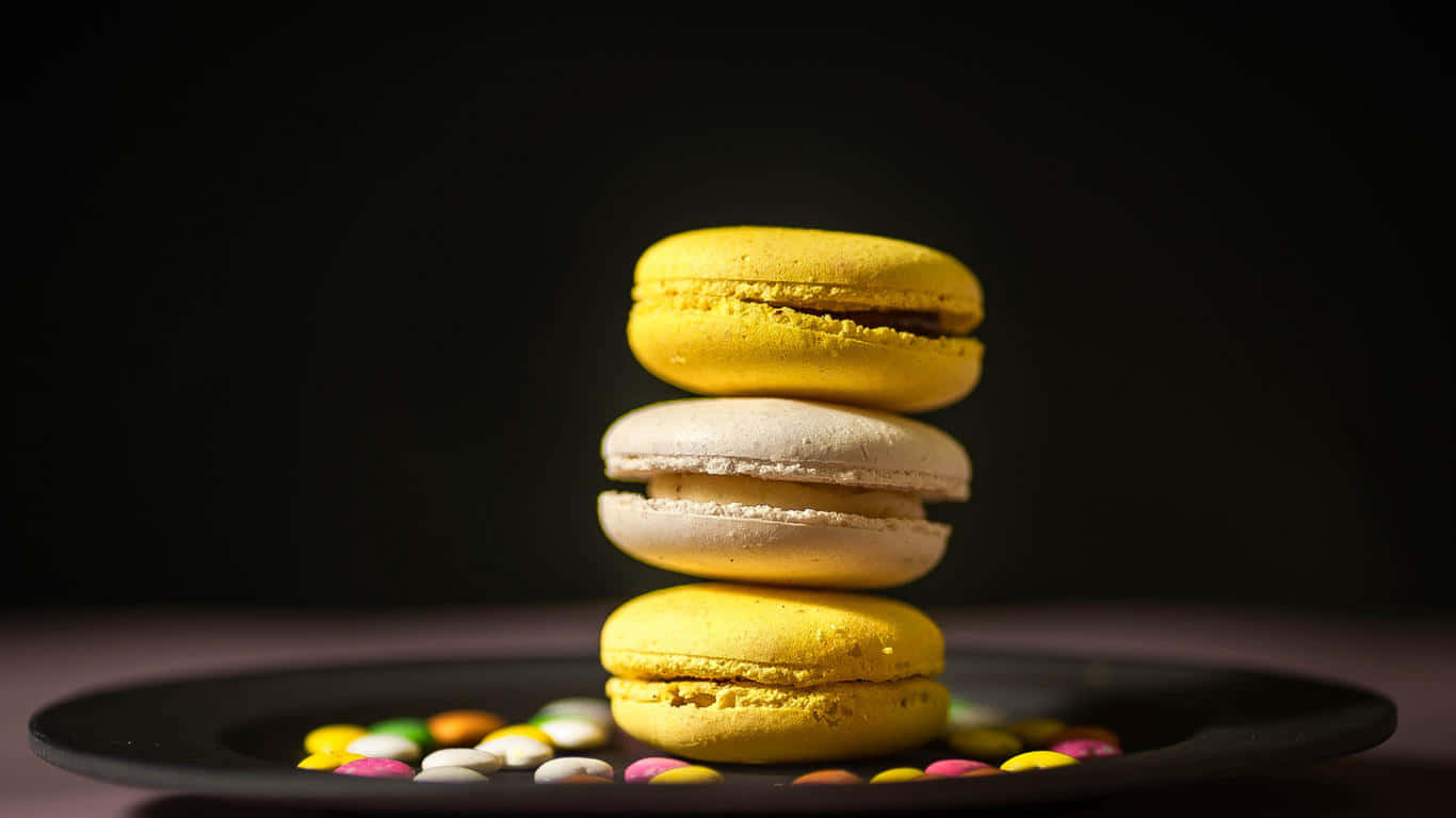1366x768 Desserts 桌布 壁紙 — 三個色彩繽紛的馬卡龍堆疊在深色盤子上，周圍散落著糖果碎片，背景為黑色。
