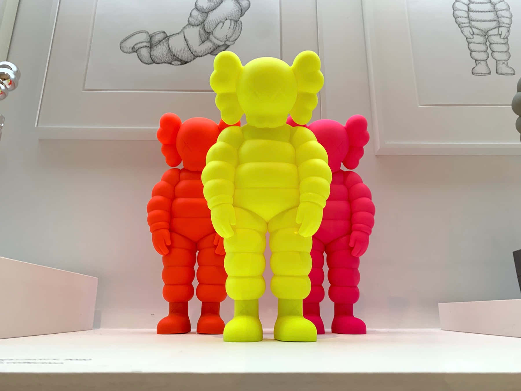 Kaws Figures 桌布 壁紙 — 三個色彩鮮豔的KAWS毛絨公仔，分別是黃色、橙色和粉紅色，排成一排放在白色架子上，背景是極簡風格的牆壁，上面掛著裱框的素描。