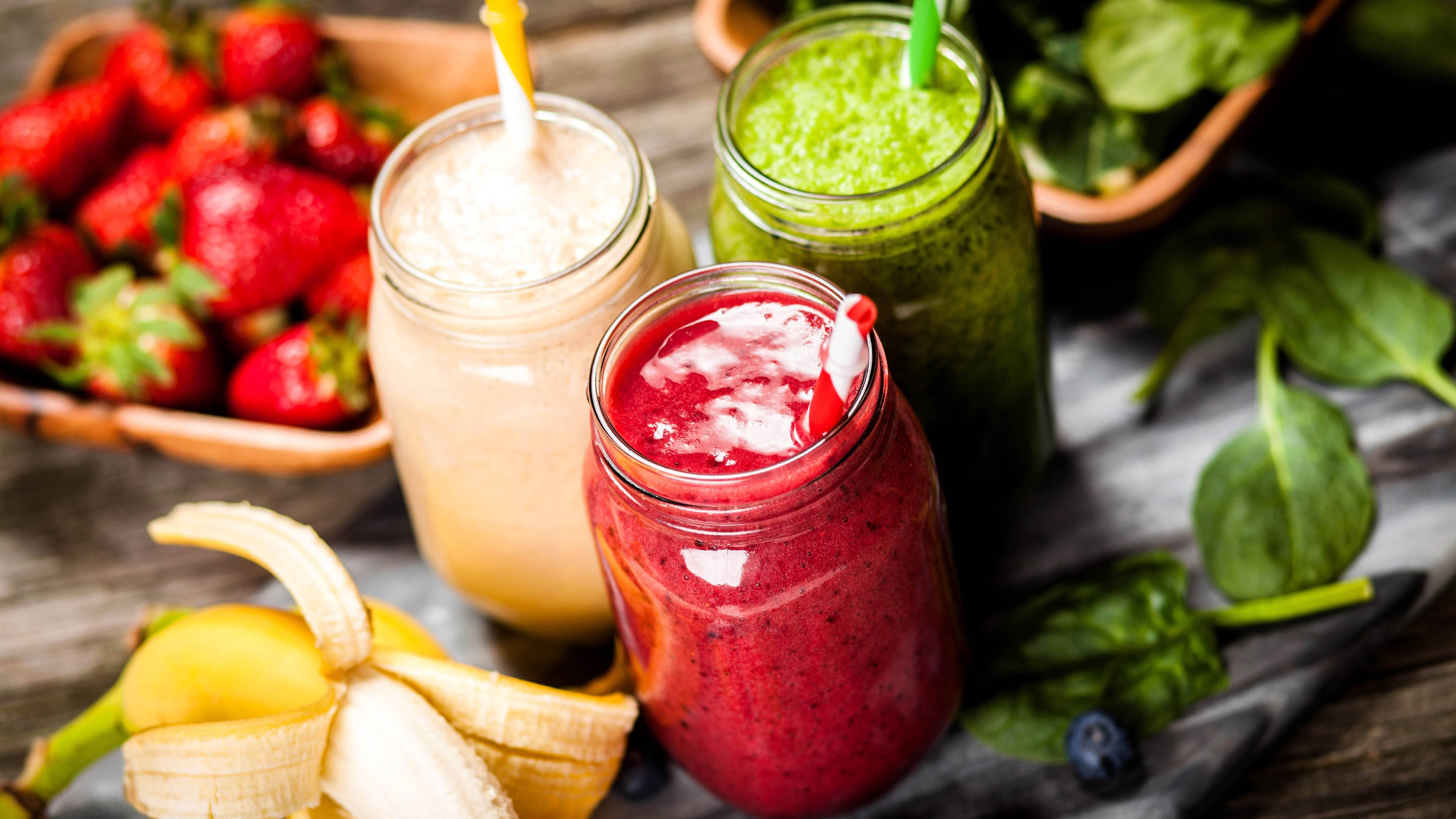 Smoothie 桌布 壁紙 — 三個色彩鮮豔的冰沙放在梅森罐中，周圍是新鮮草莓、菠菜和香蕉，置於鄉村風木桌上。