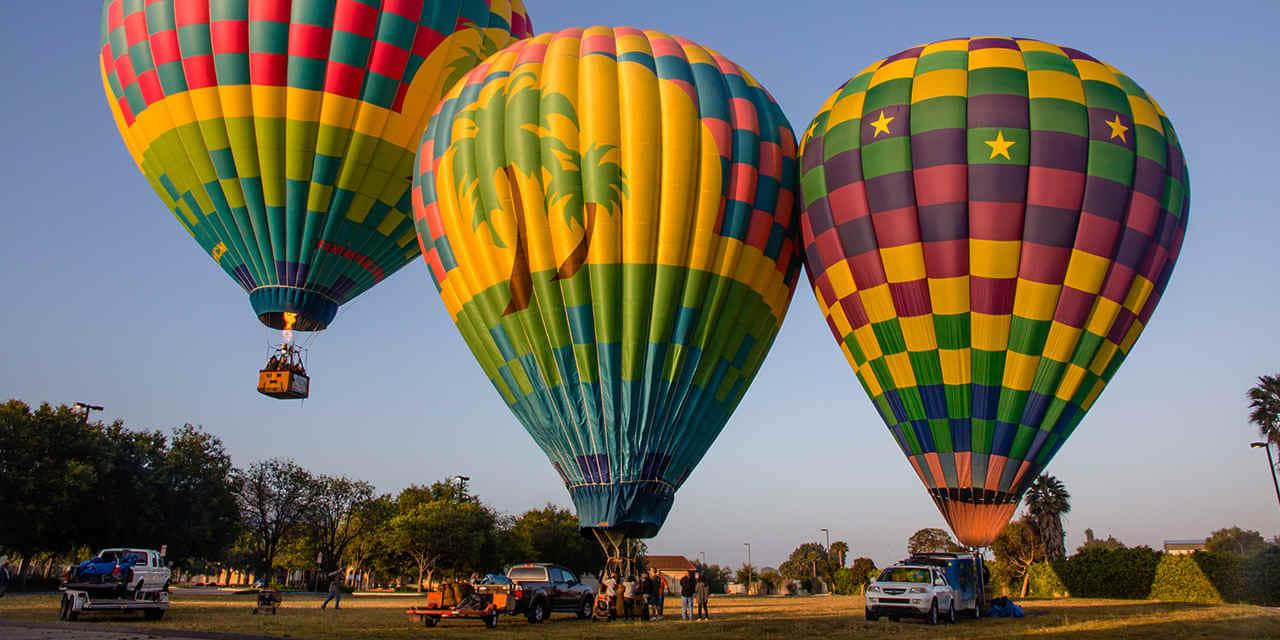 Hot Air Balloon 桌布 壁紙 — 三個色彩鮮豔的熱氣球，帶有格子和棕櫚樹圖案，在黎明時分漂浮在清澈的藍天上。
