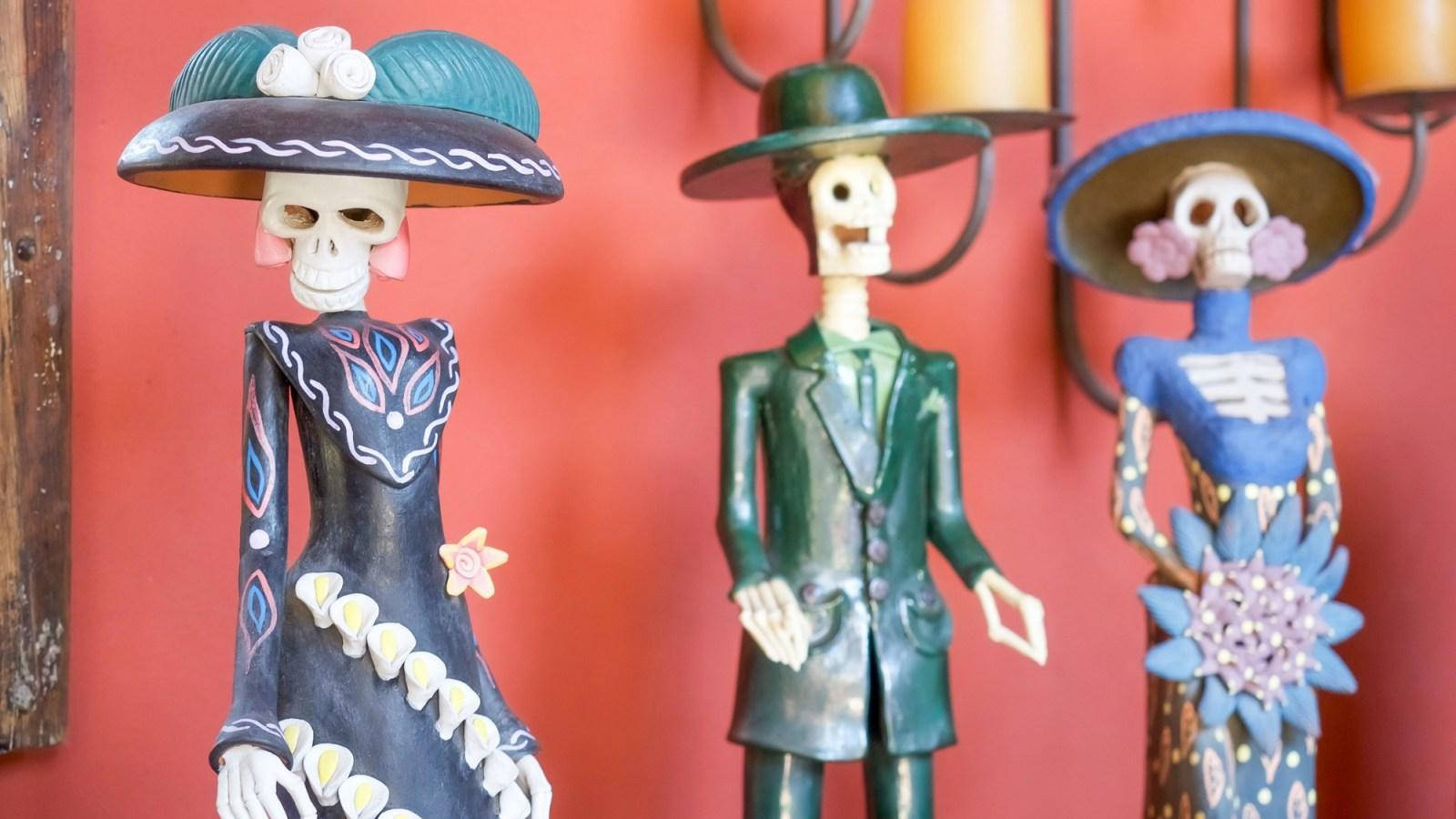Dia De Los Muertos 桌布 壁紙 — 三個色彩鮮豔的糖骷髏公仔穿著傳統服飾，站在掛有燈籠的溫暖紅牆前。