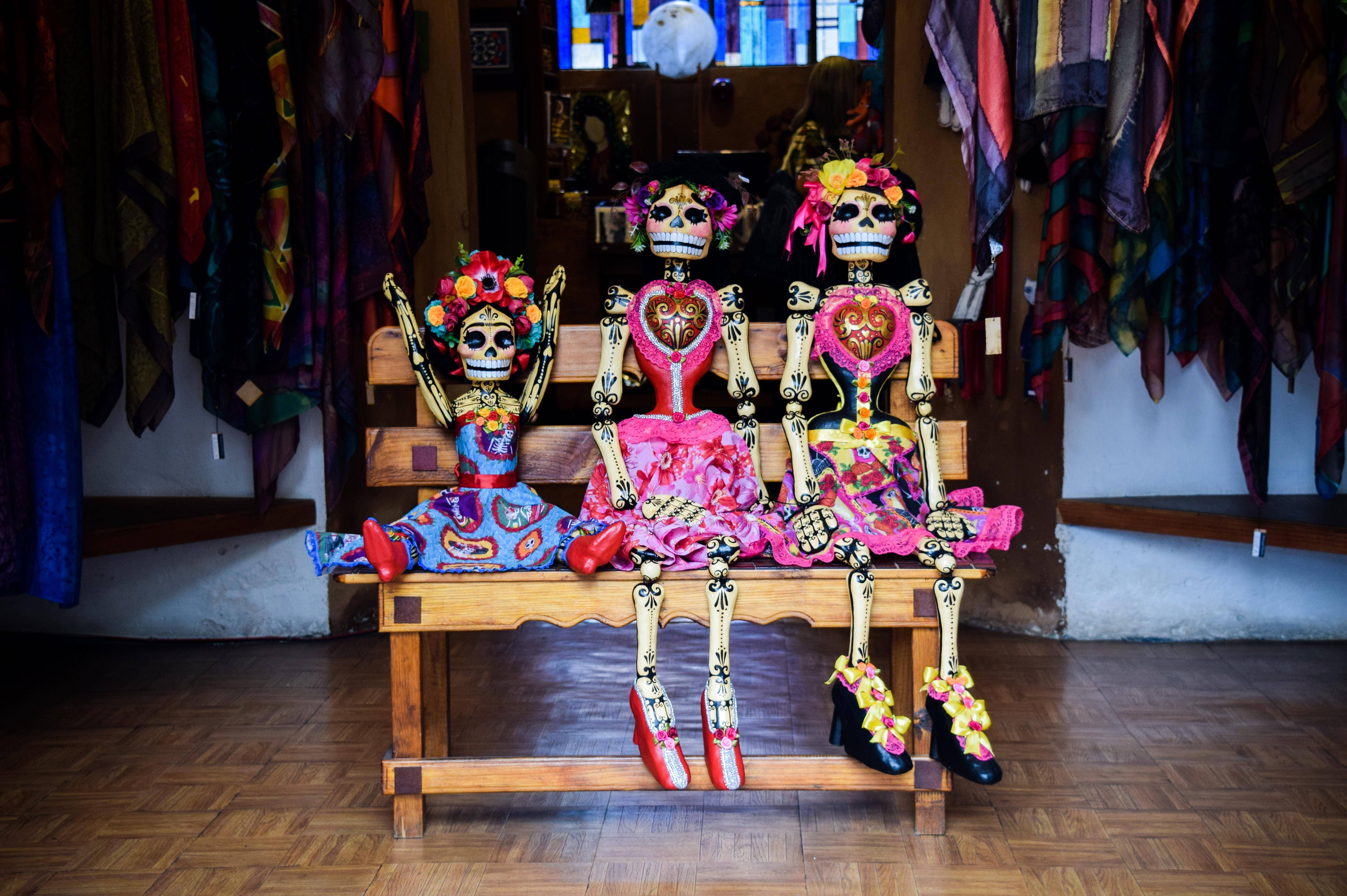 Dia De Los Muertos 桌布 壁紙 — 三個色彩鮮豔的糖骷髏娃娃坐在一間掛滿布料的彩色紡織店內的木凳上。