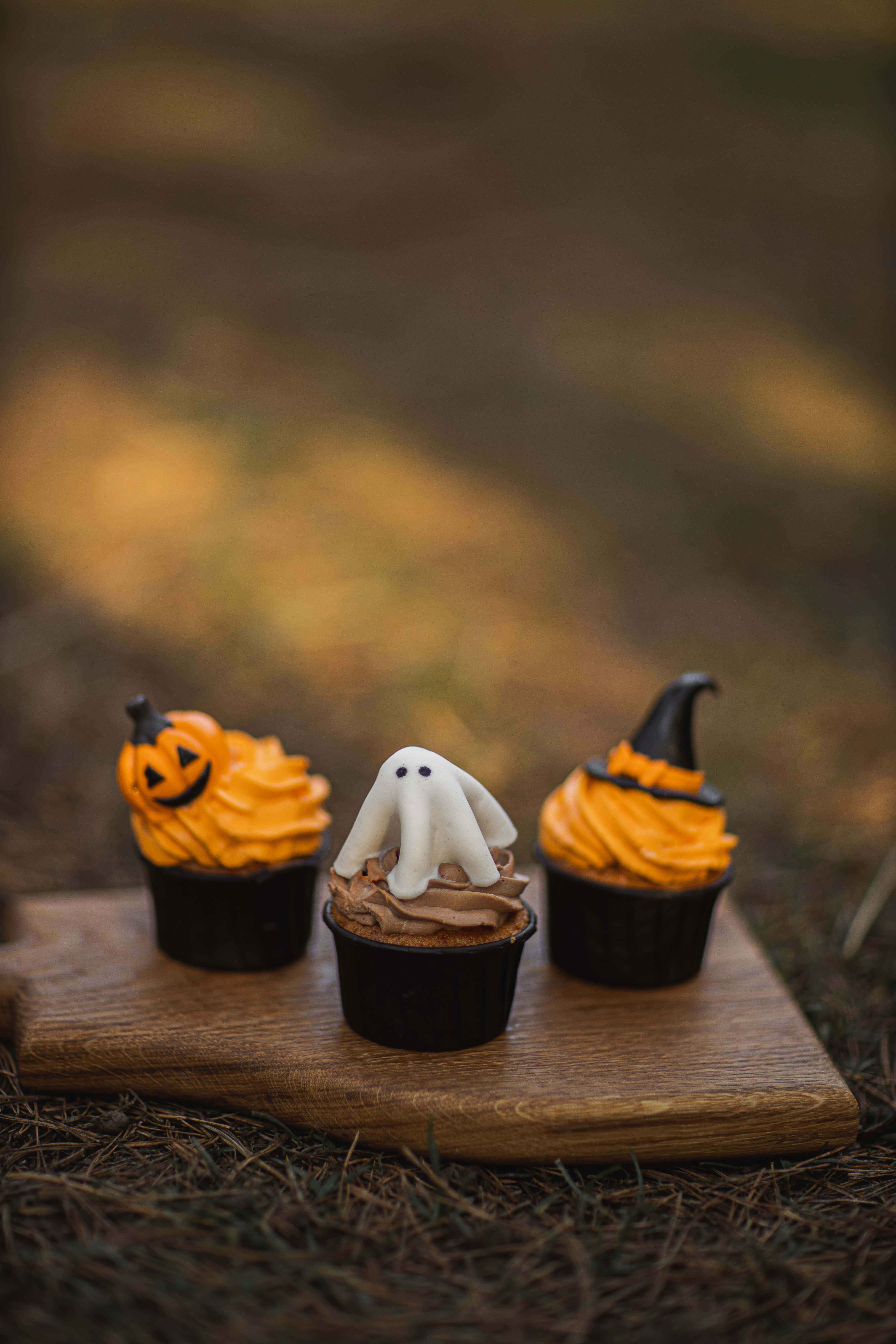 Pumpkin Patch 桌布 壁紙 — 三個萬聖節主題紙杯蛋糕，上面有南瓜燈、幽靈和女巫帽裝飾，放在戶外的木板上。