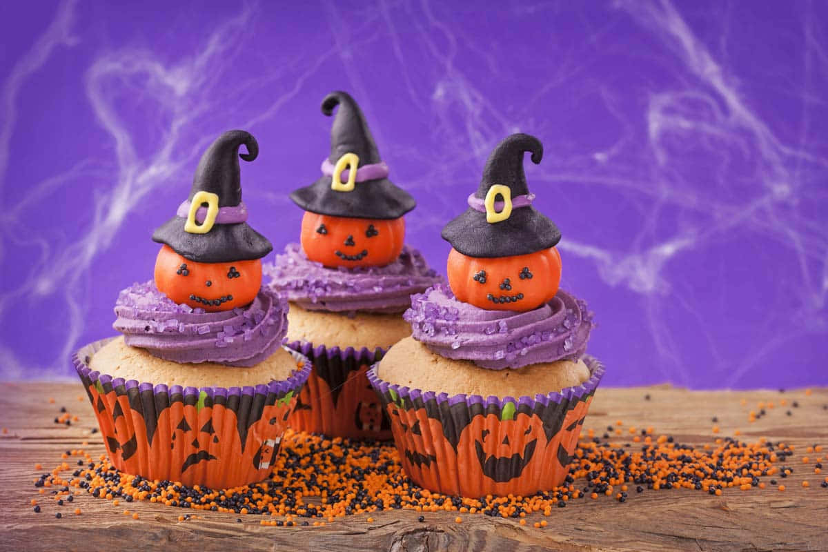 Halloween Cupcakes 桌布 壁紙 — 三個萬聖節杯子蛋糕，頂部有戴著巫婆帽的傑克南瓜燈，放在木質表面上，撒有橙色和黑色糖粒，背景是紫色蜘蛛網。