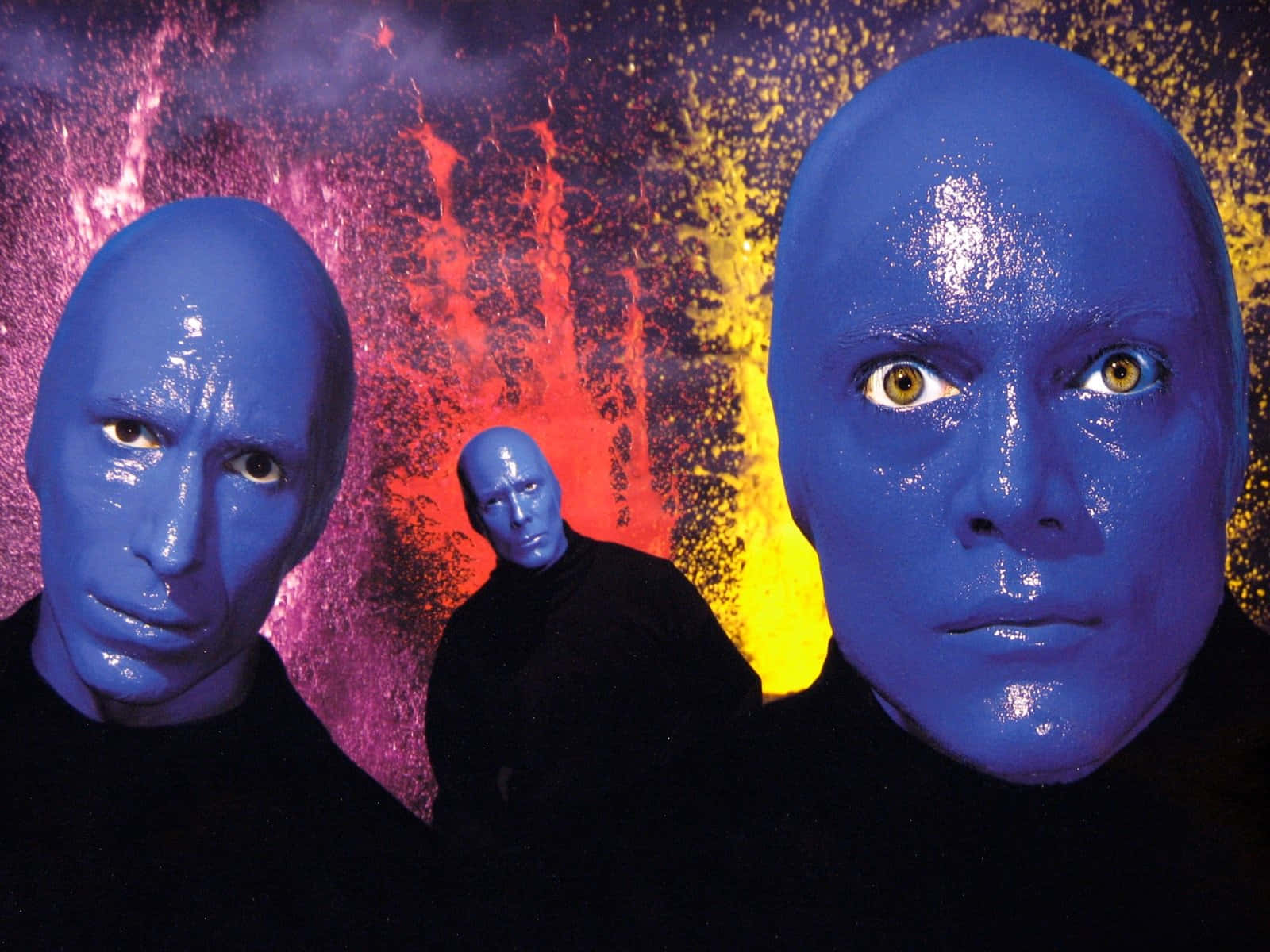 Blue Man Group 桌布 壁紙 — 三個藍皮膚、黃眼睛的人物站在充滿紅色、黃色和紫色火花的鮮豔宇宙背景前。
