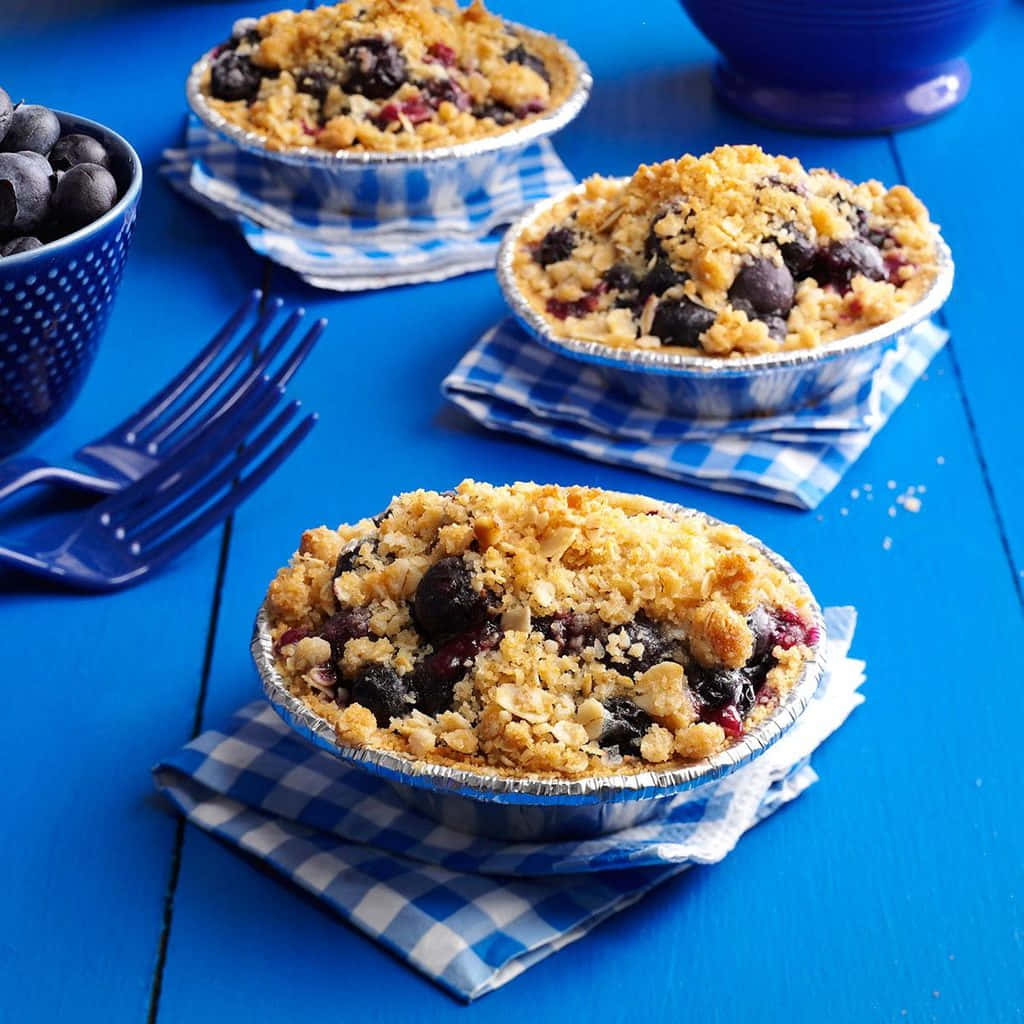 Blueberries Tart 桌布 壁紙 — 三個藍莓金寶派放在錫箔盤中，置於格紋餐巾上，再放在鮮藍色木桌上。