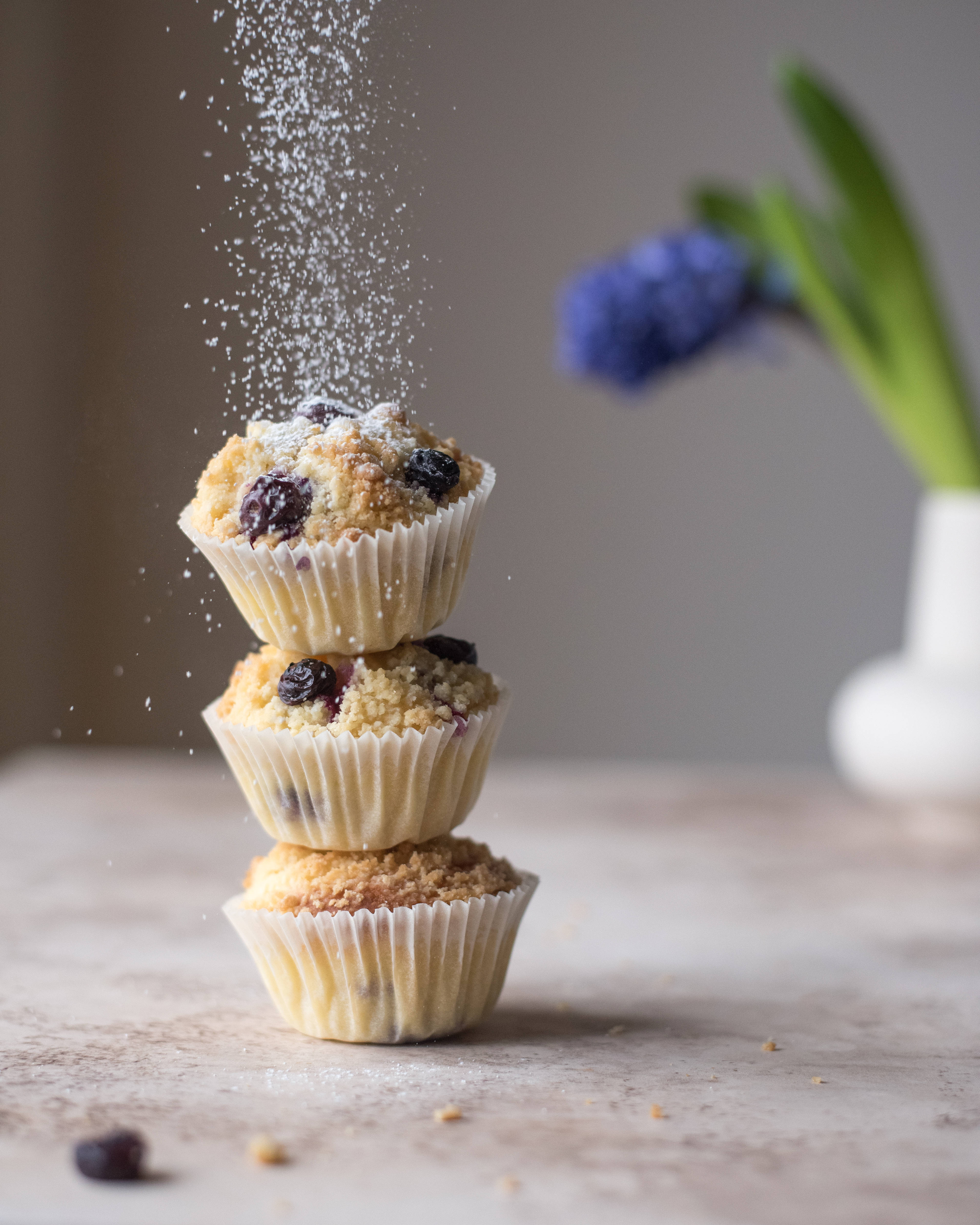 Cupcake 桌布 壁紙 — 三個藍莓馬芬疊在一起，糖粉如瀑布般流下，形成柔和模糊效果，背景柔和，配有一朵風信子。
