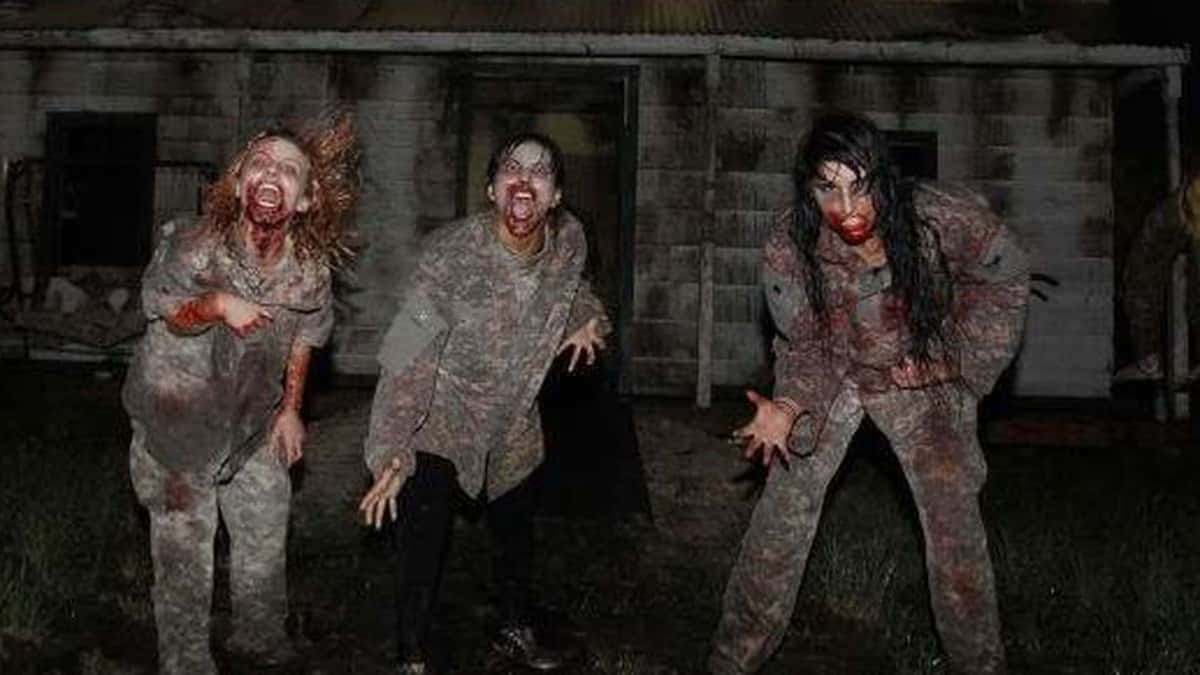 Scary Zombie 桌布 壁紙 — 三個血跡斑斑、頭髮凌亂、張開嘴巴的殭屍在夜晚一間破舊房屋前向前撲來。
