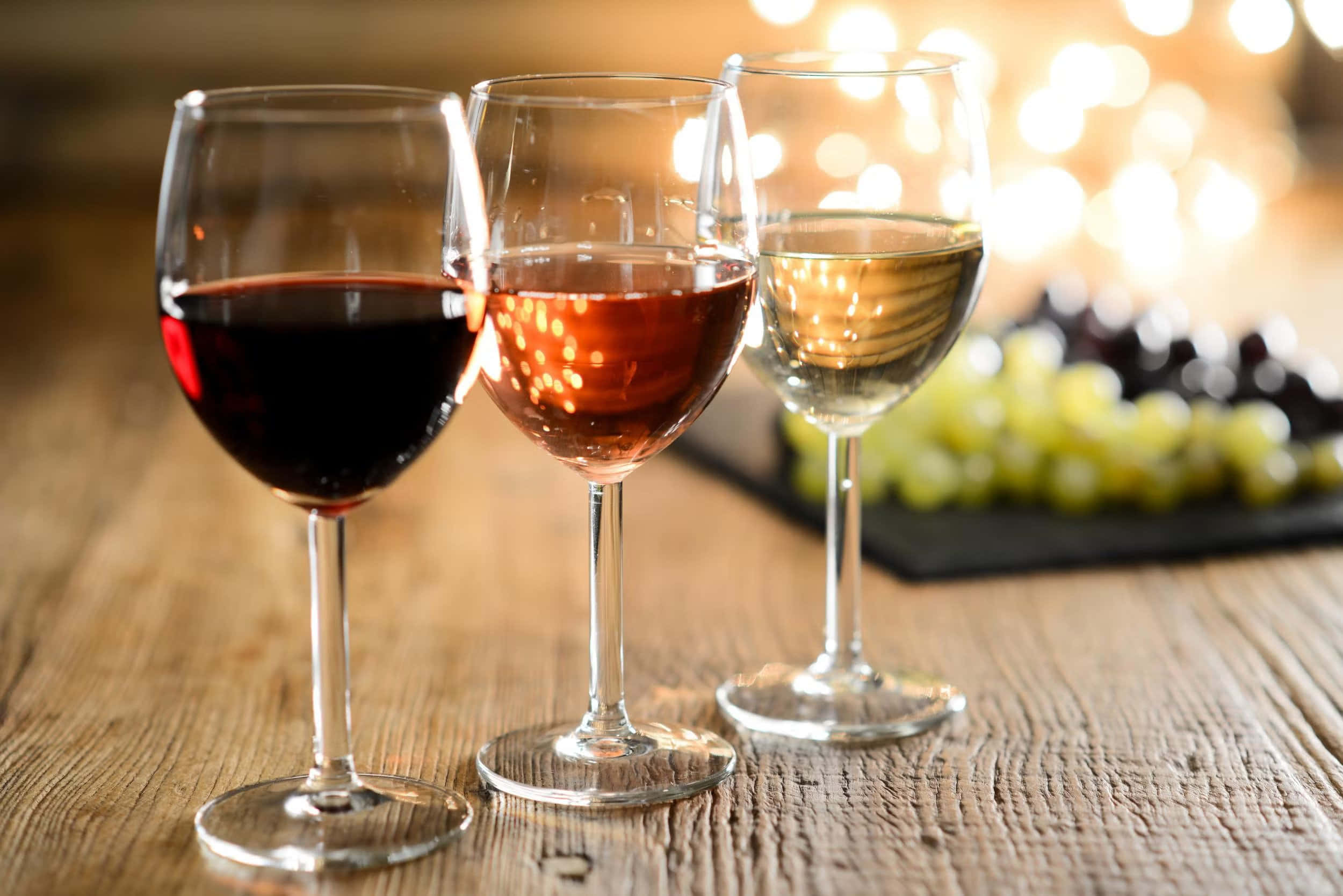 葡萄酒 桌布 壁紙 — 三個裝滿紅酒、玫瑰酒和白酒的酒杯放在一張質樸的木桌上，背景有模糊的葡萄和溫暖的散景燈光。