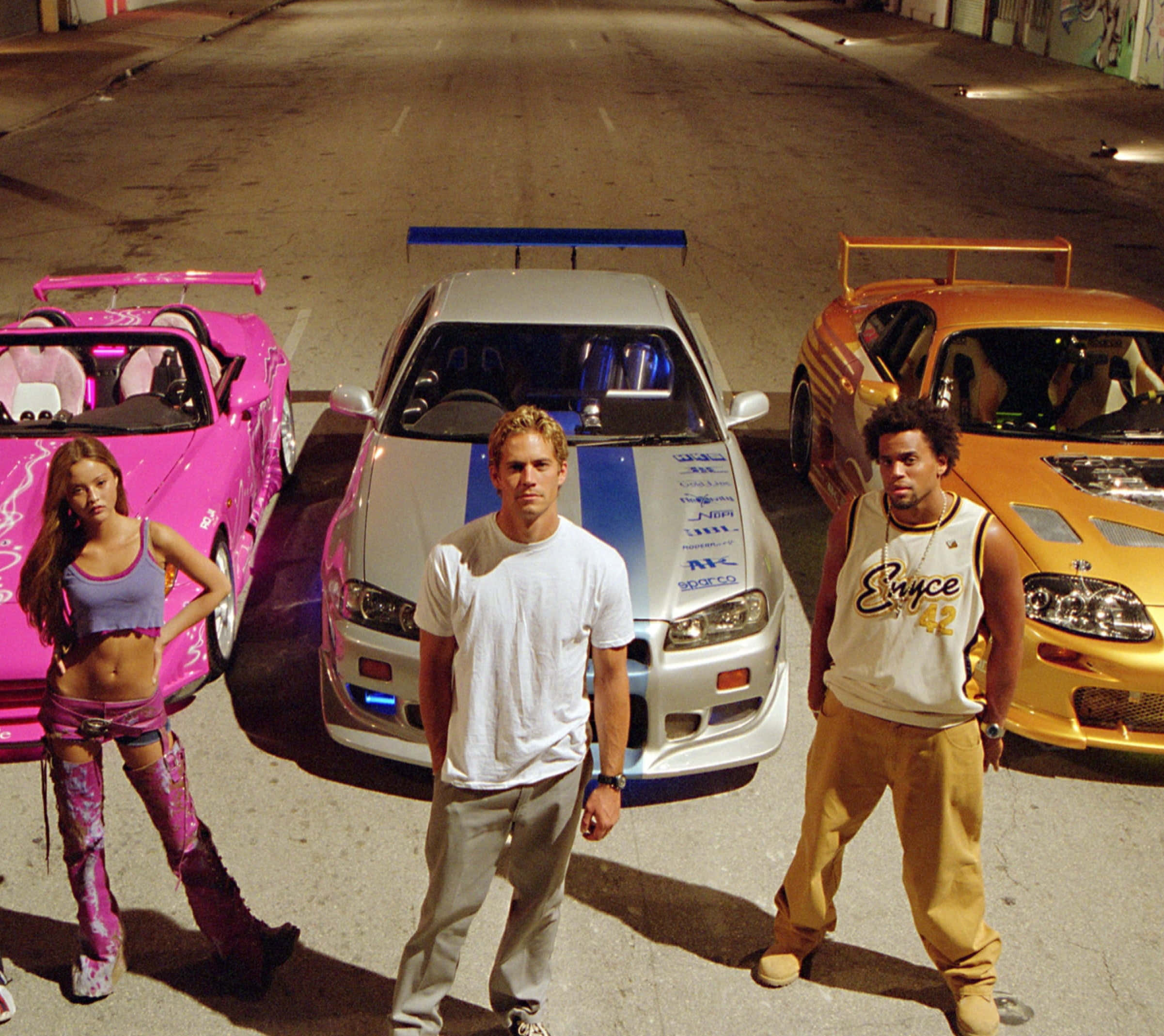 Fast And Furious 桌布 壁紙 — 三個角色自信地站在定制跑車旁邊，在光天化日之下的一條粗糙城市小巷中。