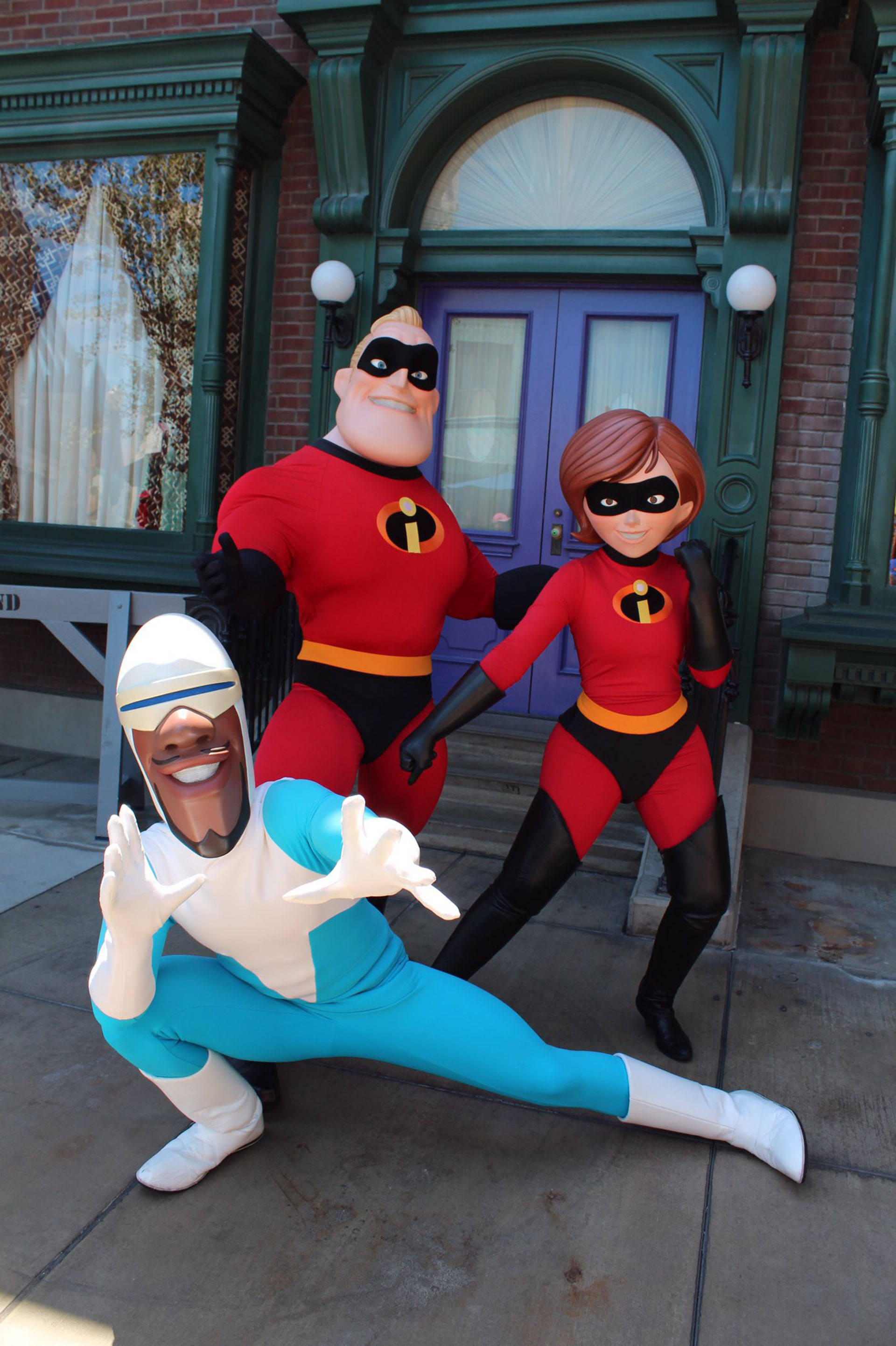 Frozone 桌布 壁紙 — 三個身穿全套服裝的超人特攻隊角色在一個有綠色飾邊和紫色門的主題建築外愉快地擺姿勢。