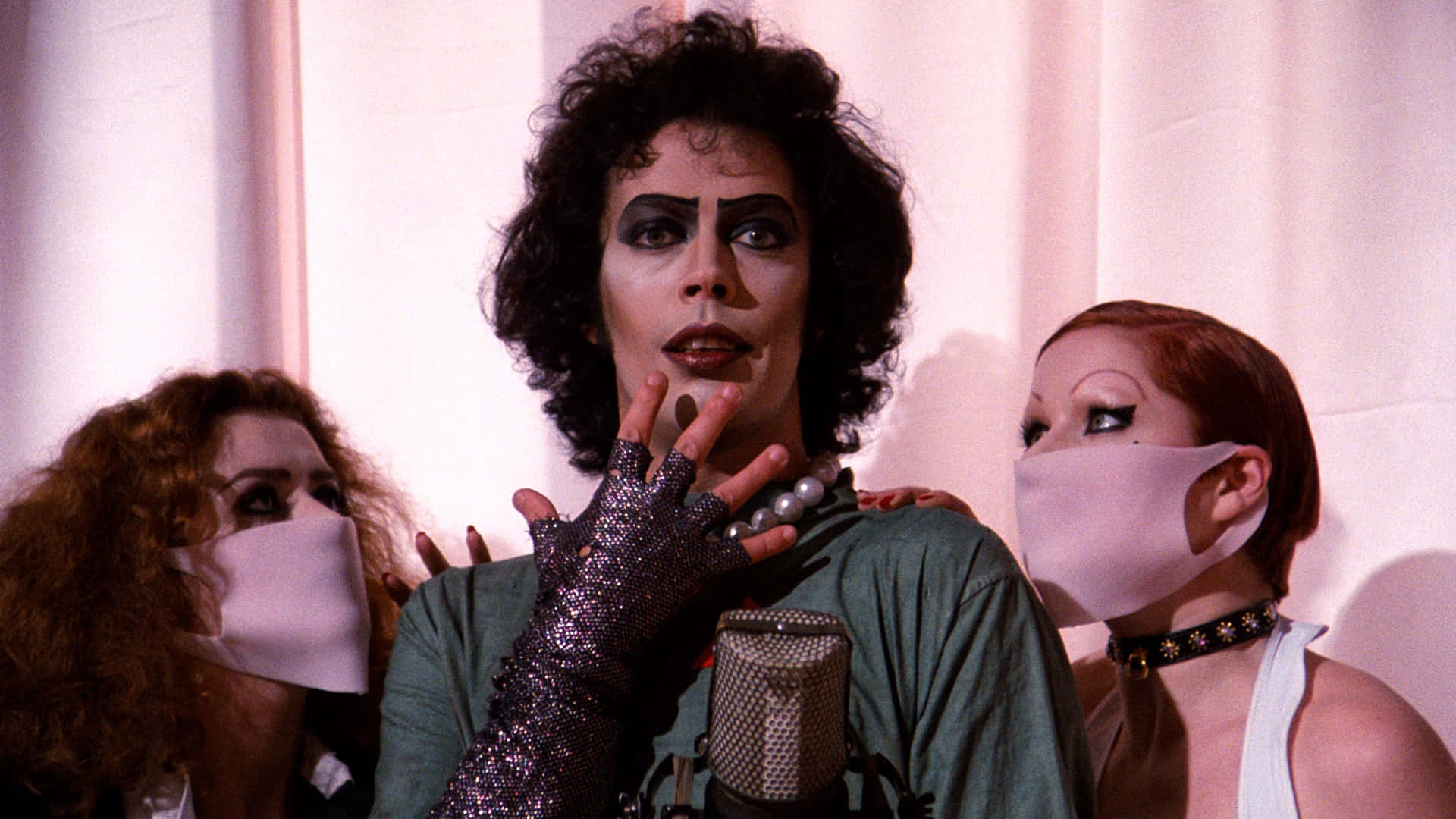 Tim Curry 桌布 壁紙 — 三個身穿戲劇化妝和面具的人物在柔軟的粉紅色窗簾前戲劇性地擺姿勢，中間的人物向一個復古麥克風做手勢。