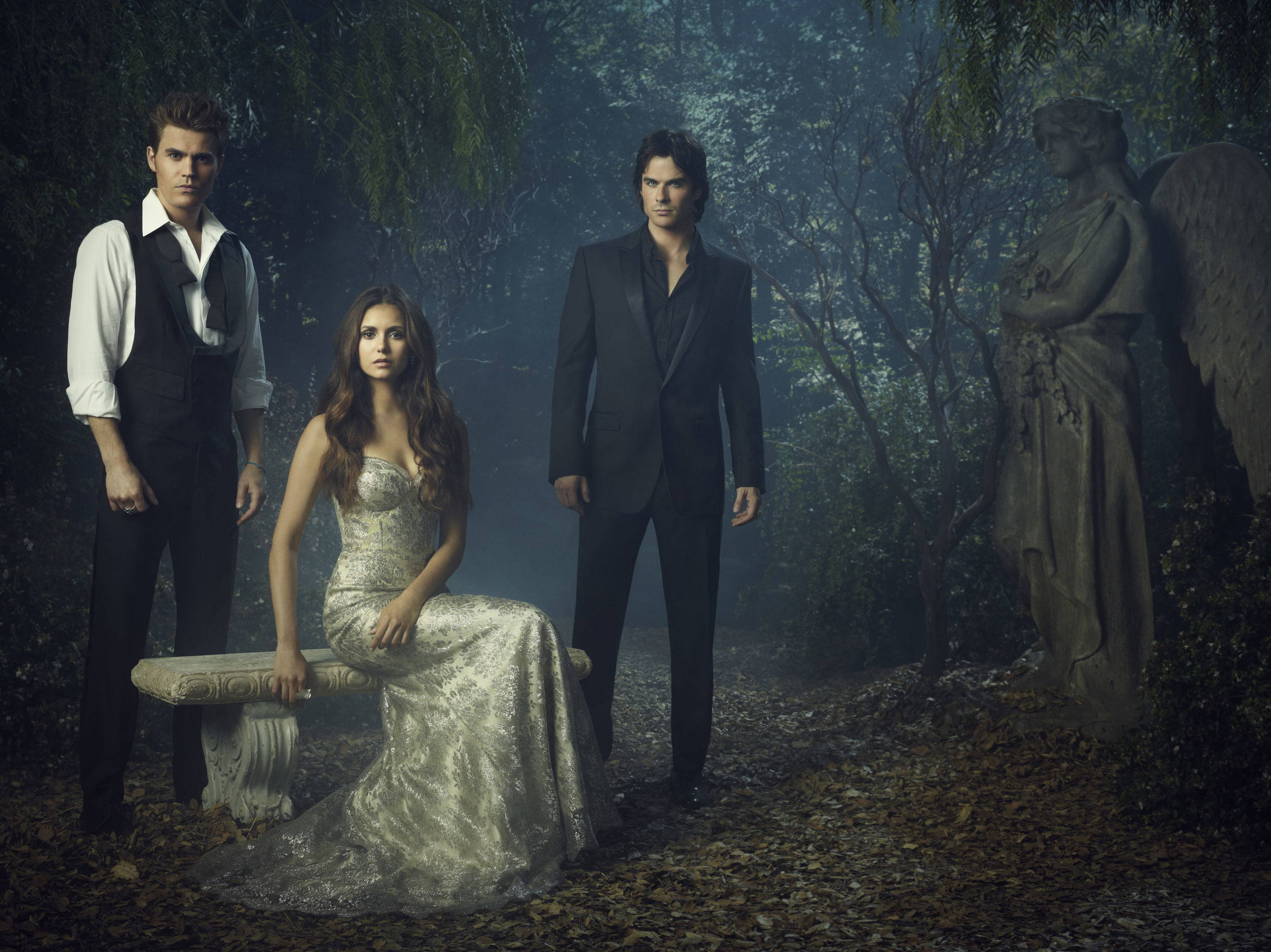 Vampire Diaries 桌布 壁紙 — 三個身穿正裝的人物站在霧氣瀰漫的森林中，旁邊有一座石天使雕像，周圍是落葉和茂密的樹木。