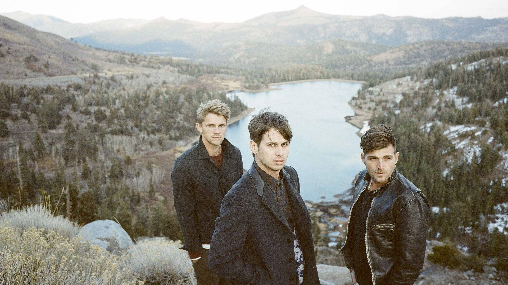foster the people 桌布 壁紙 — 三個身穿深色外套的男人站在岩石觀景點上，俯瞰著被白雪覆蓋的山脈和常綠森林環繞的蜿蜒湖泊。