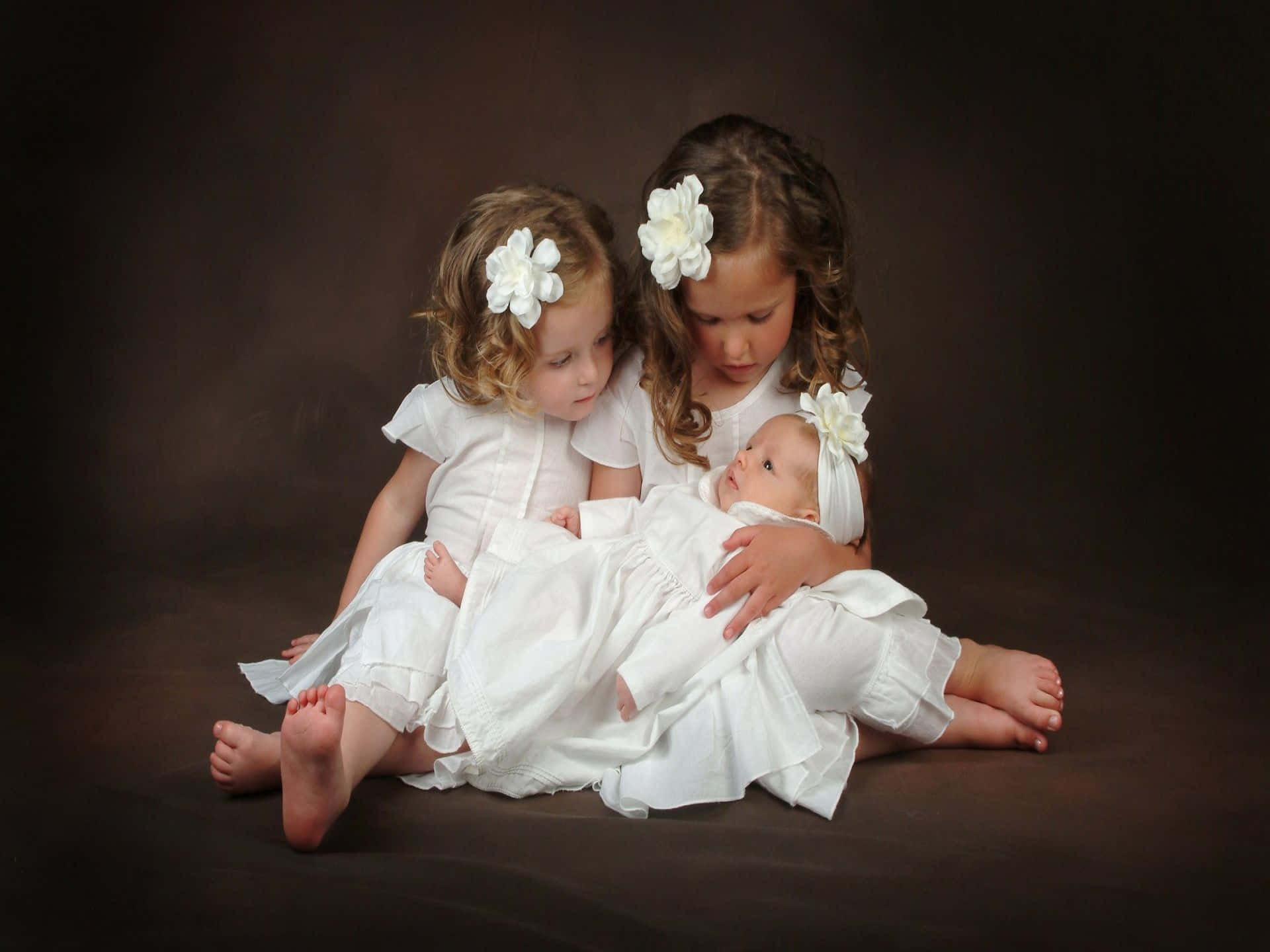 sisters 桌布 壁紙 — 三個身穿白色連衣裙、頭戴花朵髮夾的孩子緊密地坐在深棕色背景上，溫柔地抱著一個嬰兒。