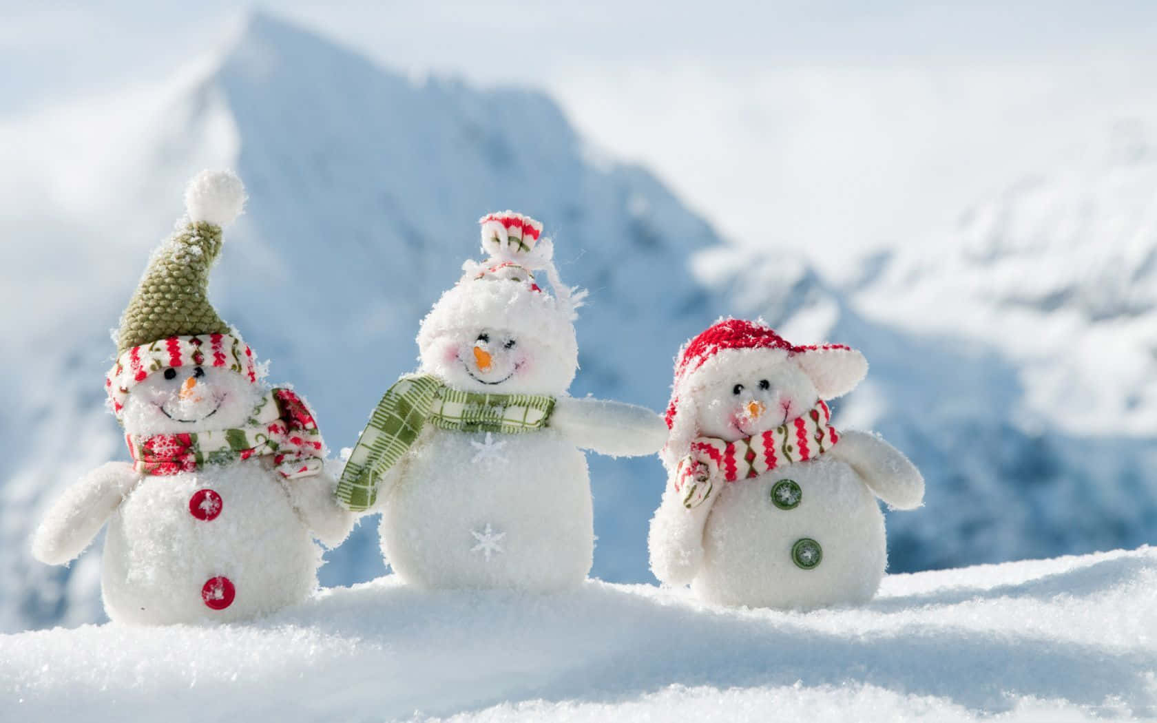  壁紙 — 三個迷人的雪人公仔戴著節日帽子和圍巾，在明亮的陽光下一起站在白雪皚皚的山坡上。