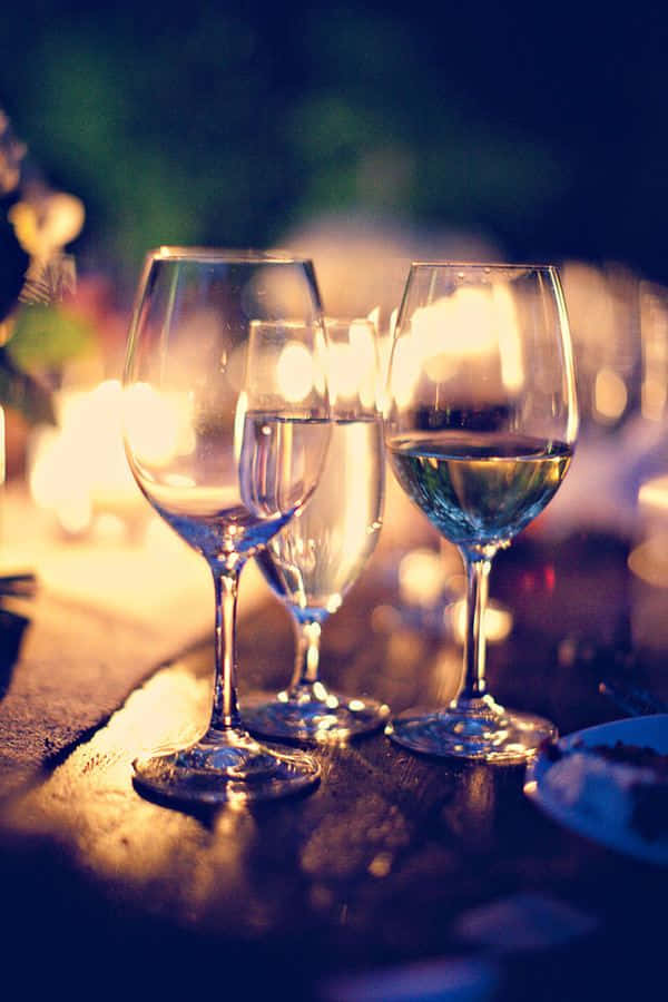 White Wine 桌布 壁紙 — 三個酒杯放在木桌上，在燭光下溫暖地閃耀，背景是模糊的花卉。