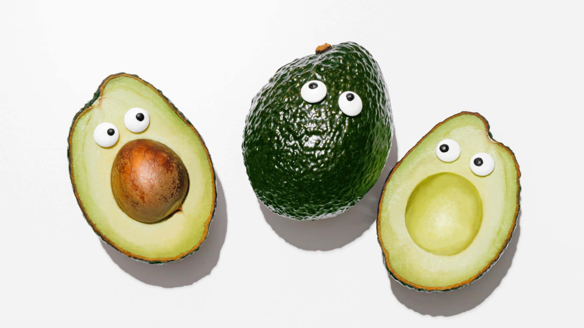 Cute Avocado 桌布 壁紙 — 三個酪梨帶有咕嚕眼睛，排列在純白色背景上，一個完整，一個切半有籽，一個切半空心。