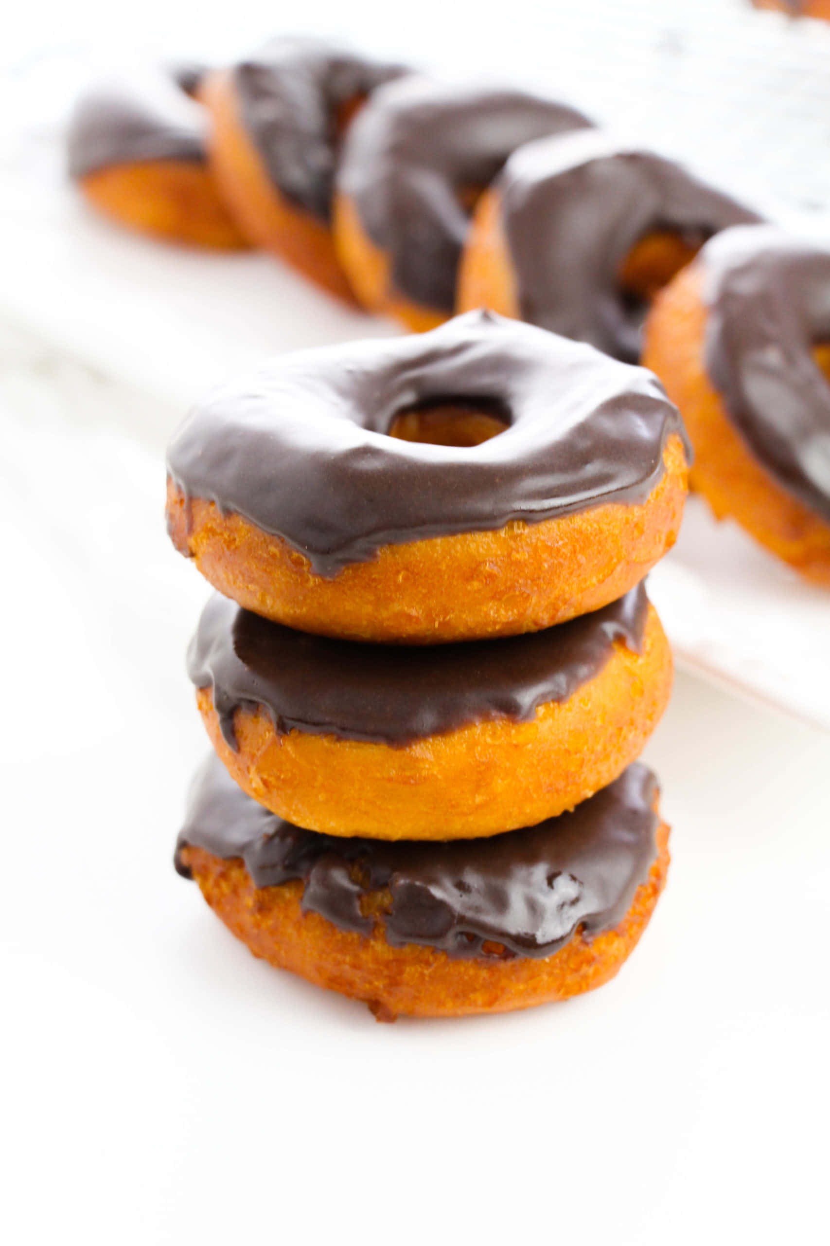 Glazed Donut 桌布 壁紙 — 三個金棕色巧克力釉面甜甜圈堆疊在焦點中，背景有更多模糊的甜甜圈在白色表面上。