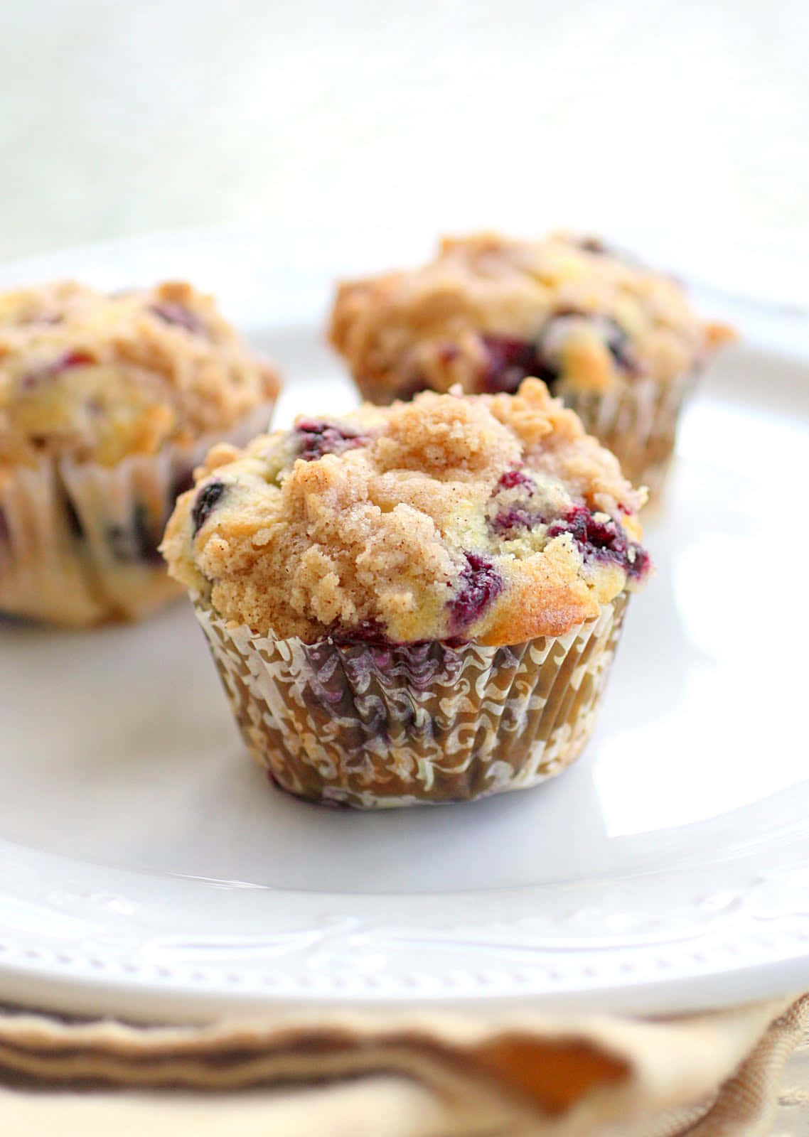 Blueberry Muffins 桌布 壁紙 — 三個金黃色藍莓馬芬，上面有酥脆的奶酥粒，放在白色盤子上，配以米色餐巾。