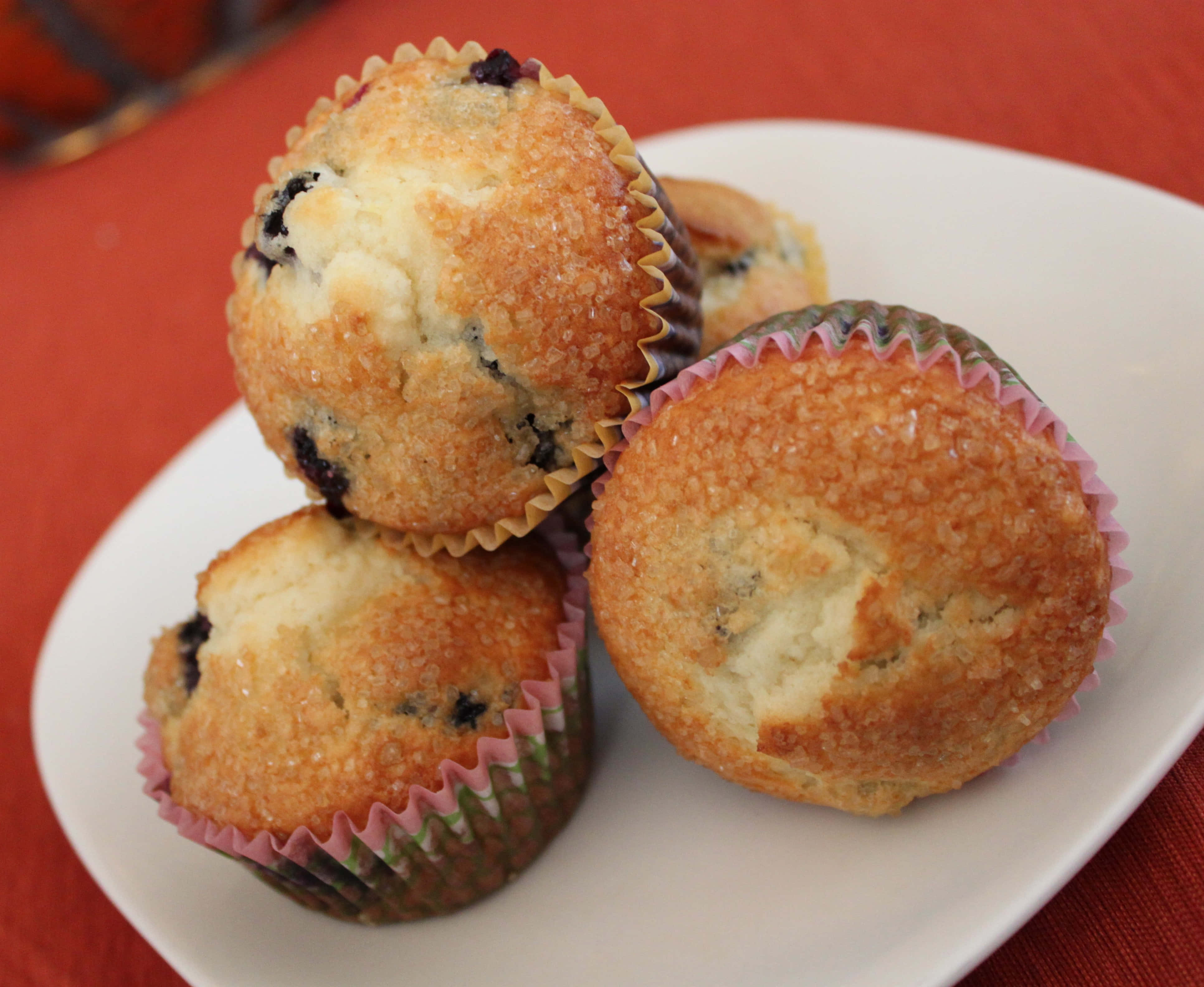 Blueberry Muffins 桌布 壁紙 — 三個金黃色藍莓馬芬放在彩色紙托中，堆疊在白色盤子上，背景為紅色布料。
