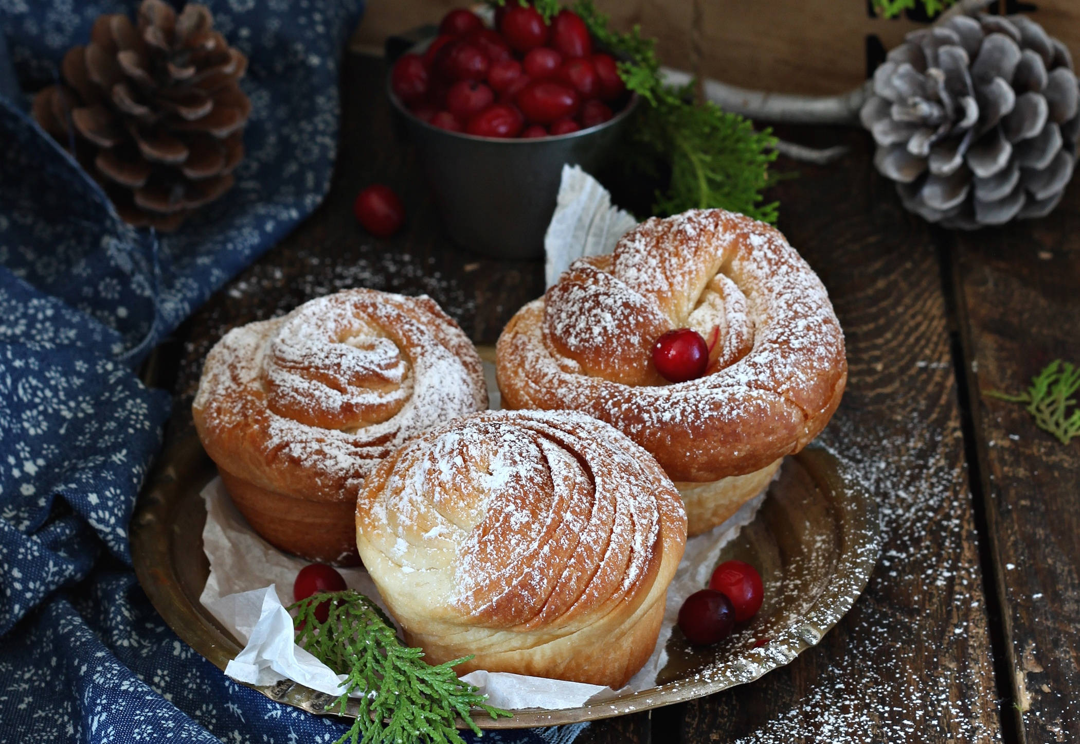 Pastries 桌布 壁紙 — 三個金黃色辮子狀糕點撒上糖粉，放在鄉村風格托盤上，旁邊有蔓越莓、松果和常綠枝條。