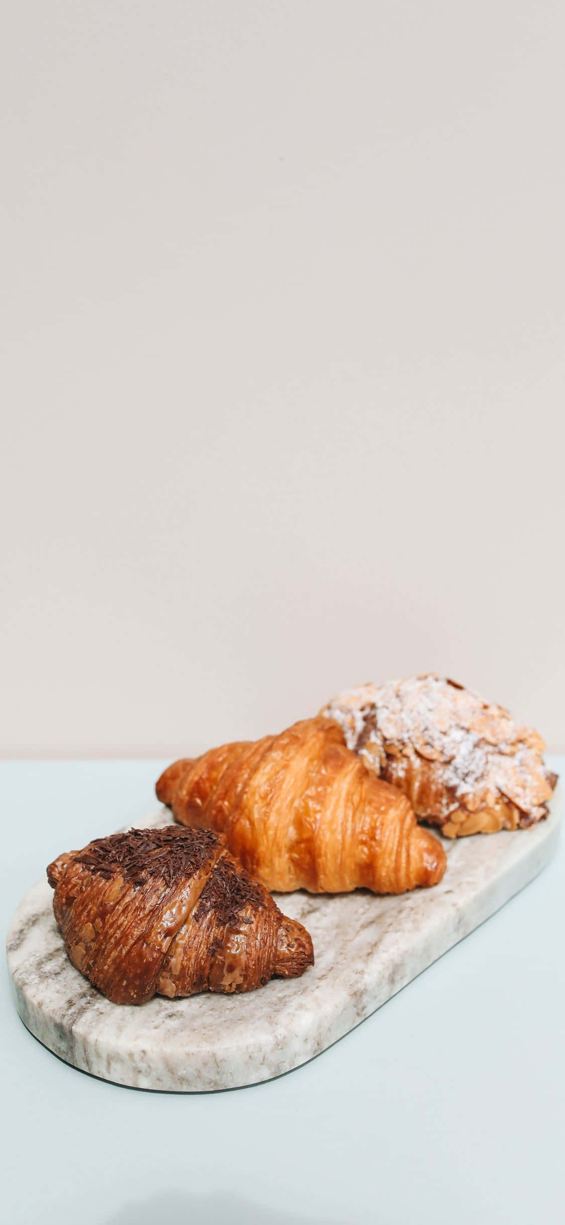 Pastry 桌布 壁紙 — 三個金黃酥脆、撒上糖粉的可頌，優雅地放在白色大理石板上面，襯托柔和粉彩背景。