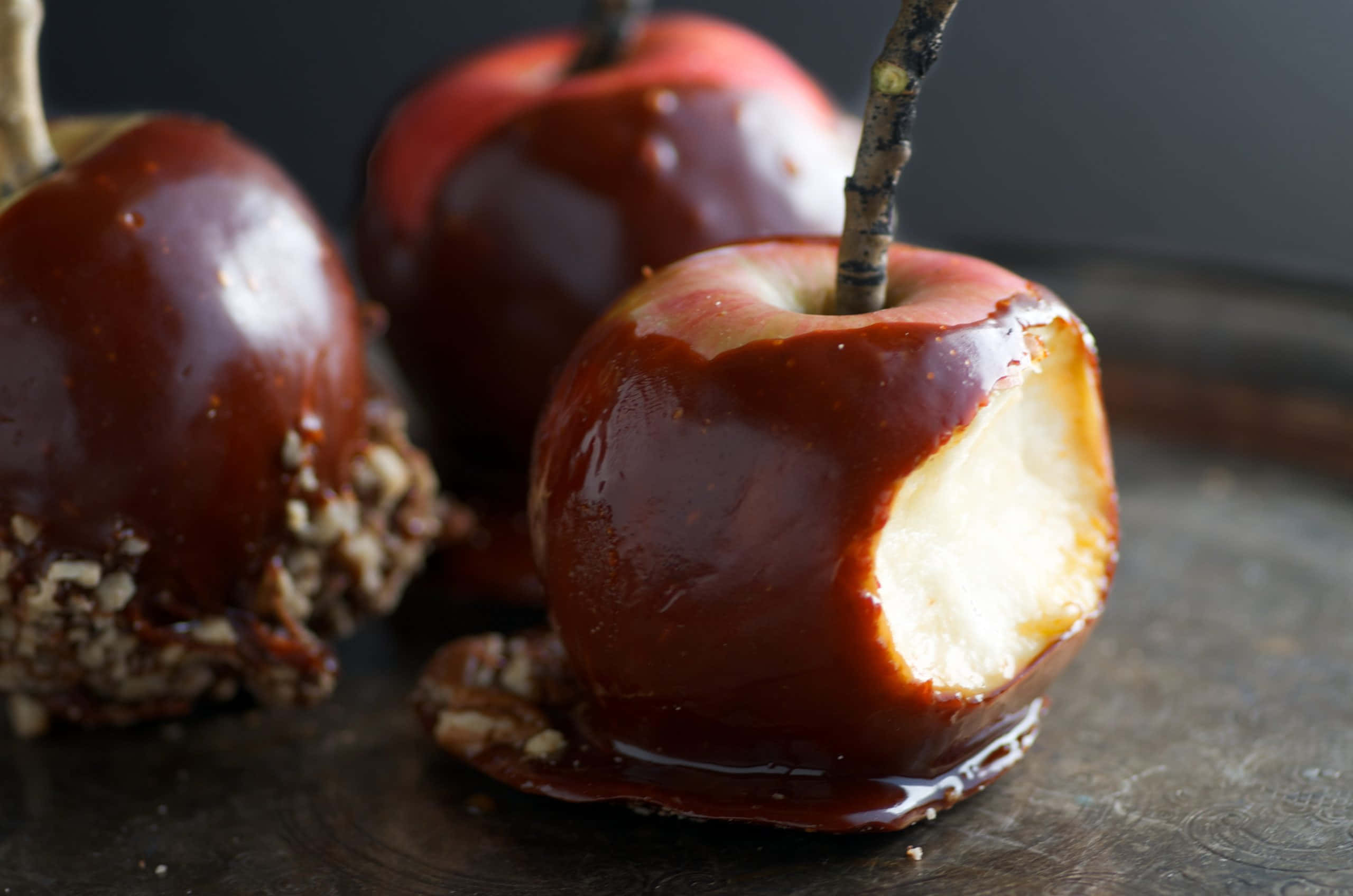 Caramel Apples 桌布 壁紙 — 三個閃亮的焦糖蘋果，其中一個被咬了一口，放在一個深色托盤上，其中一個裹著碎堅果。
