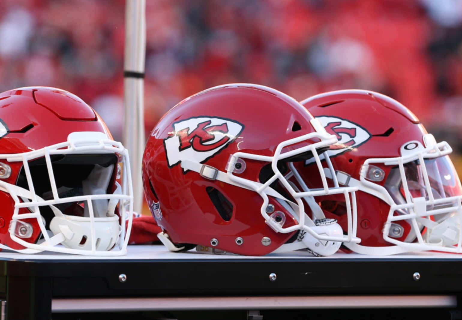 Kansas City Chiefs 4K 桌布 壁紙 — 三個閃亮的紅色堪薩斯城酋長隊美式足球頭盔，帶有白色面罩，放在黑色長椅上，背景是模糊的體育場人群。
