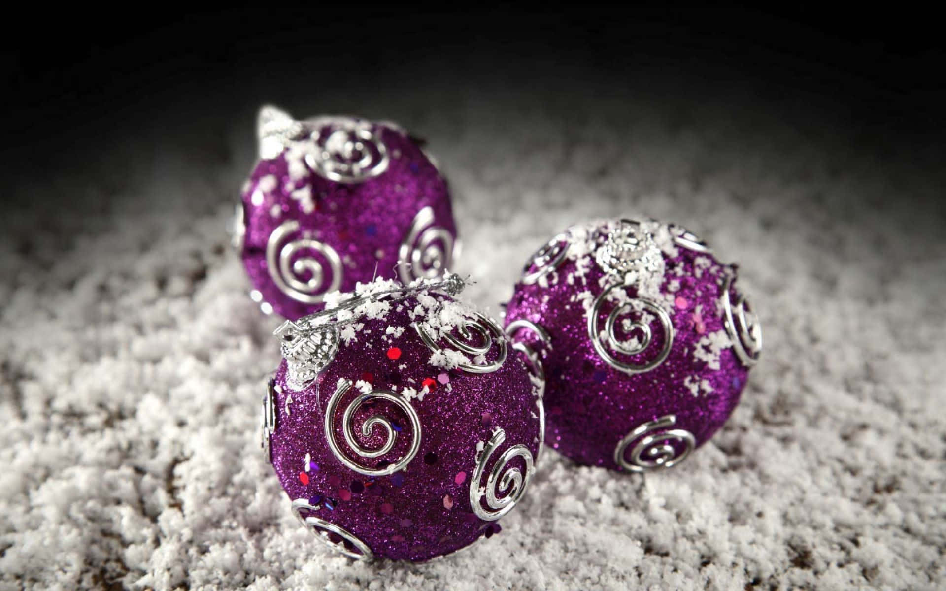 Purple Christmas 桌布 壁紙 — 三個閃亮紫色聖誕裝飾品，帶有銀色漩渦，放在白色雪地上，背景深色。