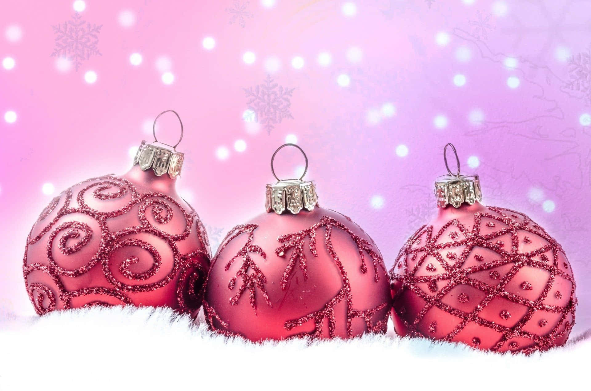 pink christmas 桌布 壁紙 — 三個閃閃發光的粉紅色聖誕裝飾品，帶有精緻圖案，放在蓬鬆的白雪上，背景是柔和的粉紅色，帶有散景燈光和雪花圖案。
