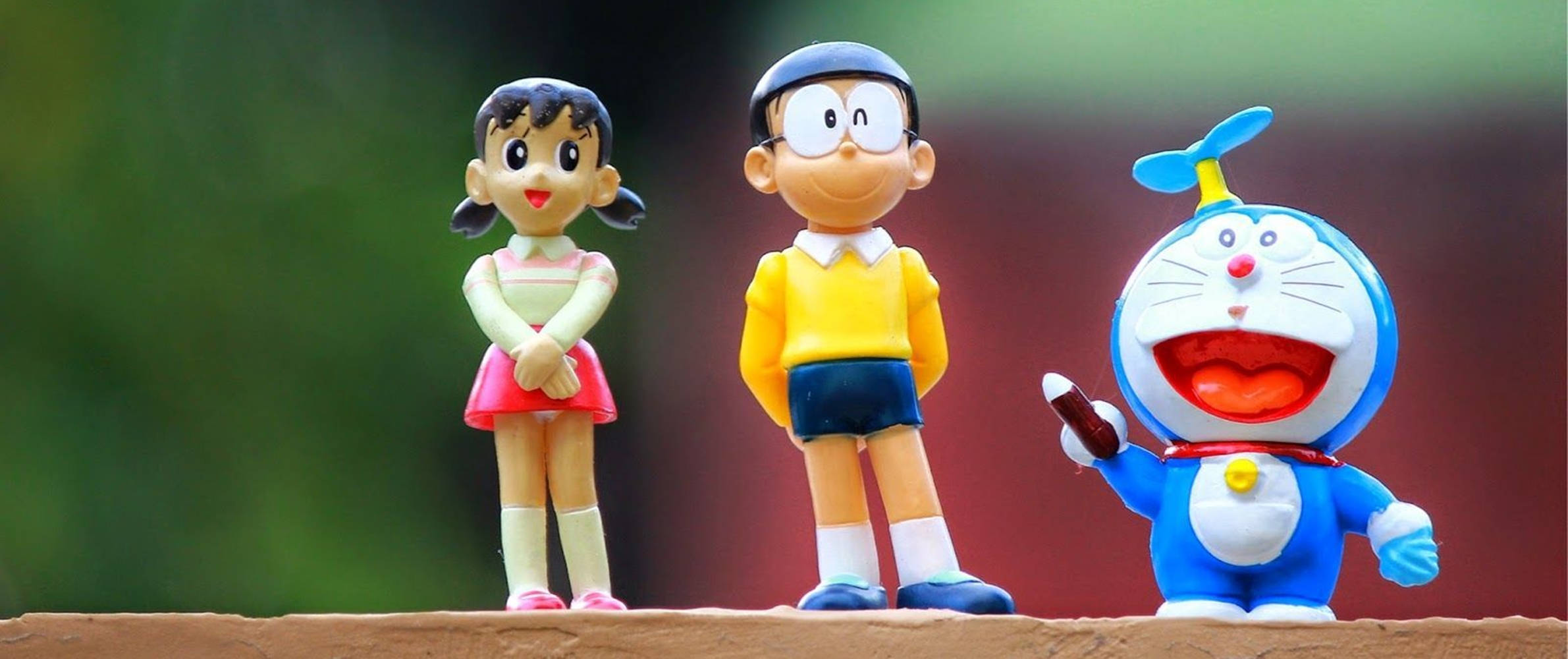 nobita 桌布 壁紙 — 三個開心的哆啦A夢動畫公仔一起站在木質表面上，背景是模糊的綠色和紅色。