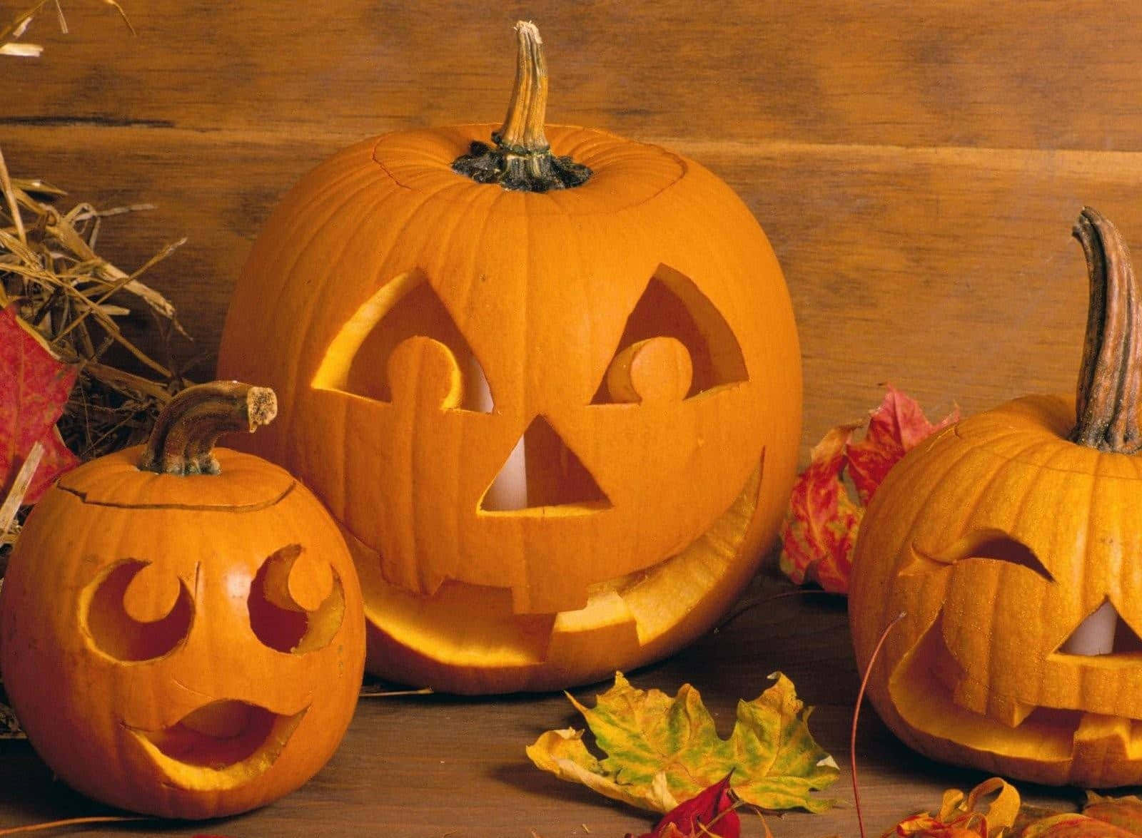 Pumpkin 桌布 壁紙 — 三個雕刻的南瓜燈籠，發光的臉孔，放在木桌上，周圍有秋葉和乾草。