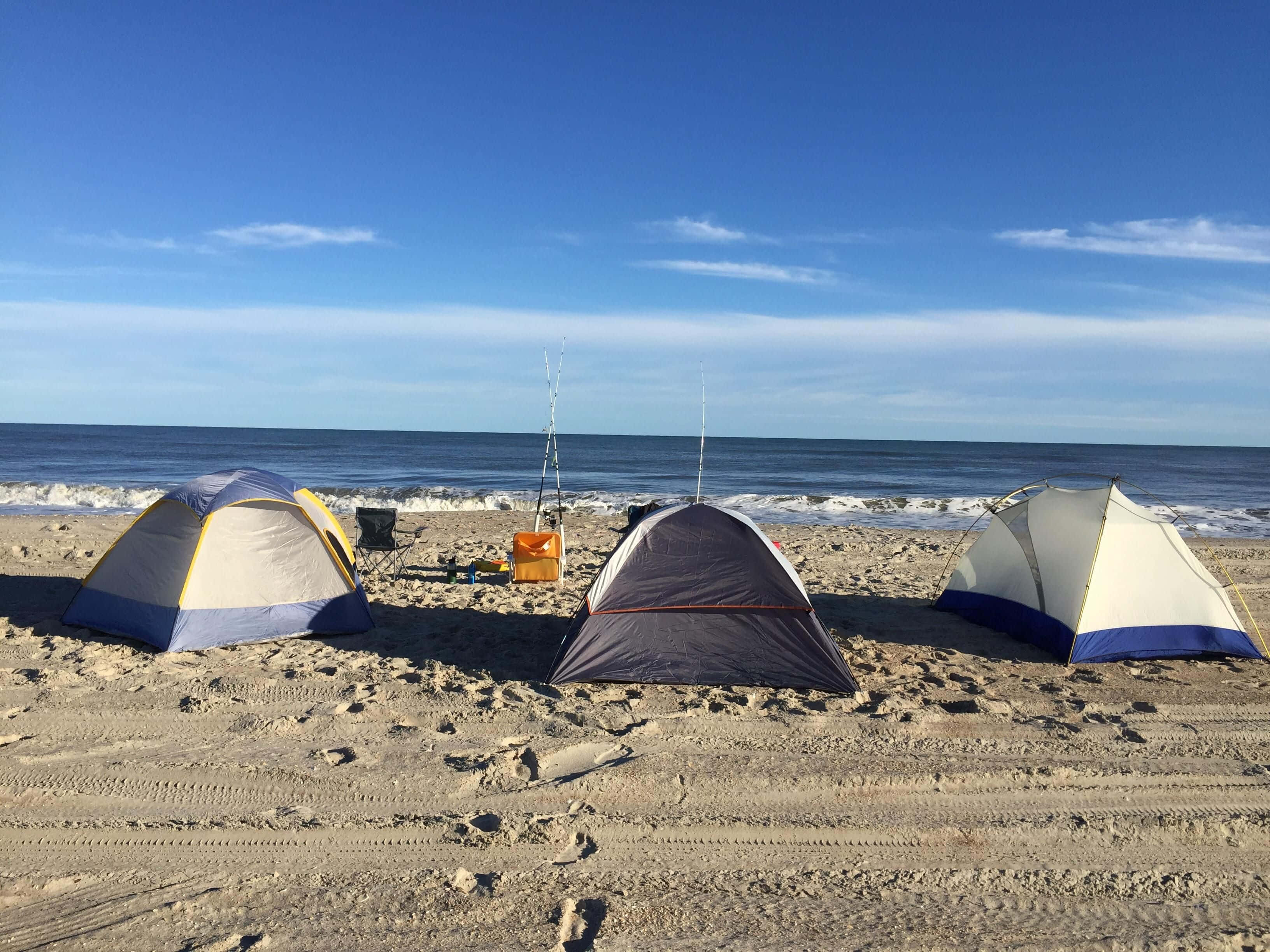 Beach Camping 桌布 壁紙 — 三個露營帳篷設置在沙灘上，有海浪和晴朗的藍天。